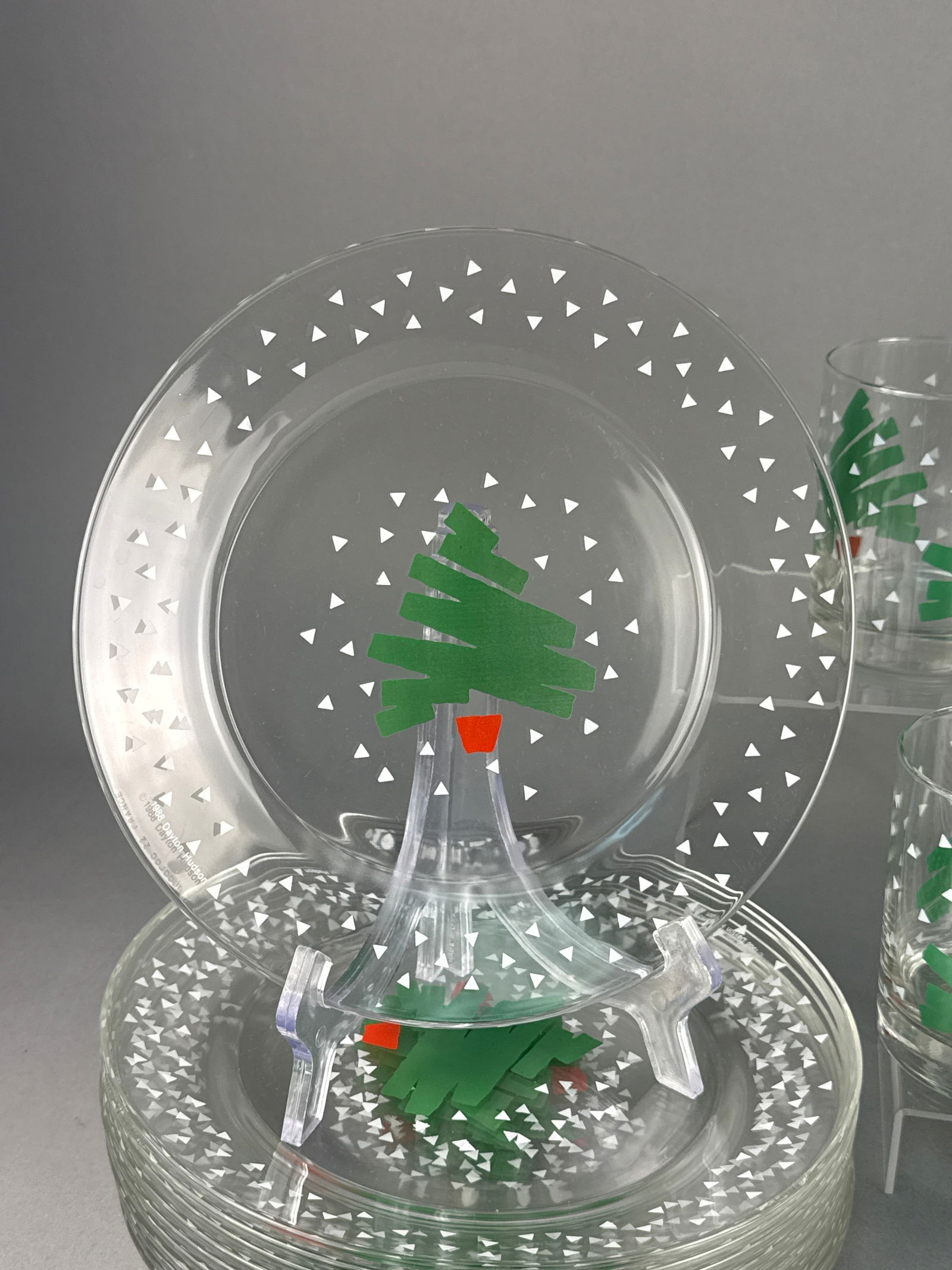 A Collection of Dayton Hudson Arcoroc Tree & Confetti Pattern Glassware: A collection of clear glass holiday-themed tableware in the Tree & Confetti pattern, manufactured by Arcoroc for Dayton Hudson. The set consists of circular plates and cylindrical tumblers. Each p