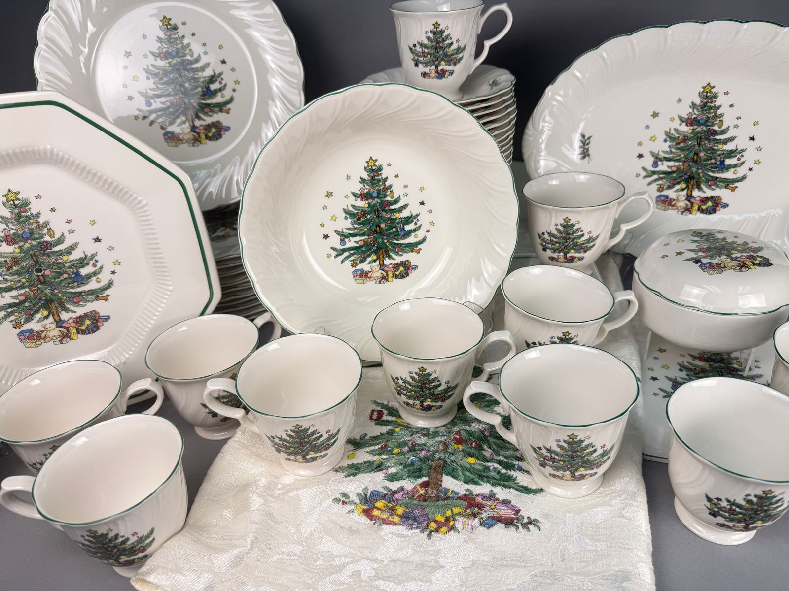A Collection of Nikko Happy Holidays Ceramic Tableware and a Spode Tablecloth: A collection of ceramic tableware in the Happy Holidays pattern by Nikko, Japan, accompanied by a Spode brand tablecloth. The ceramic items feature a Christmas tree motif decorated with ornaments and