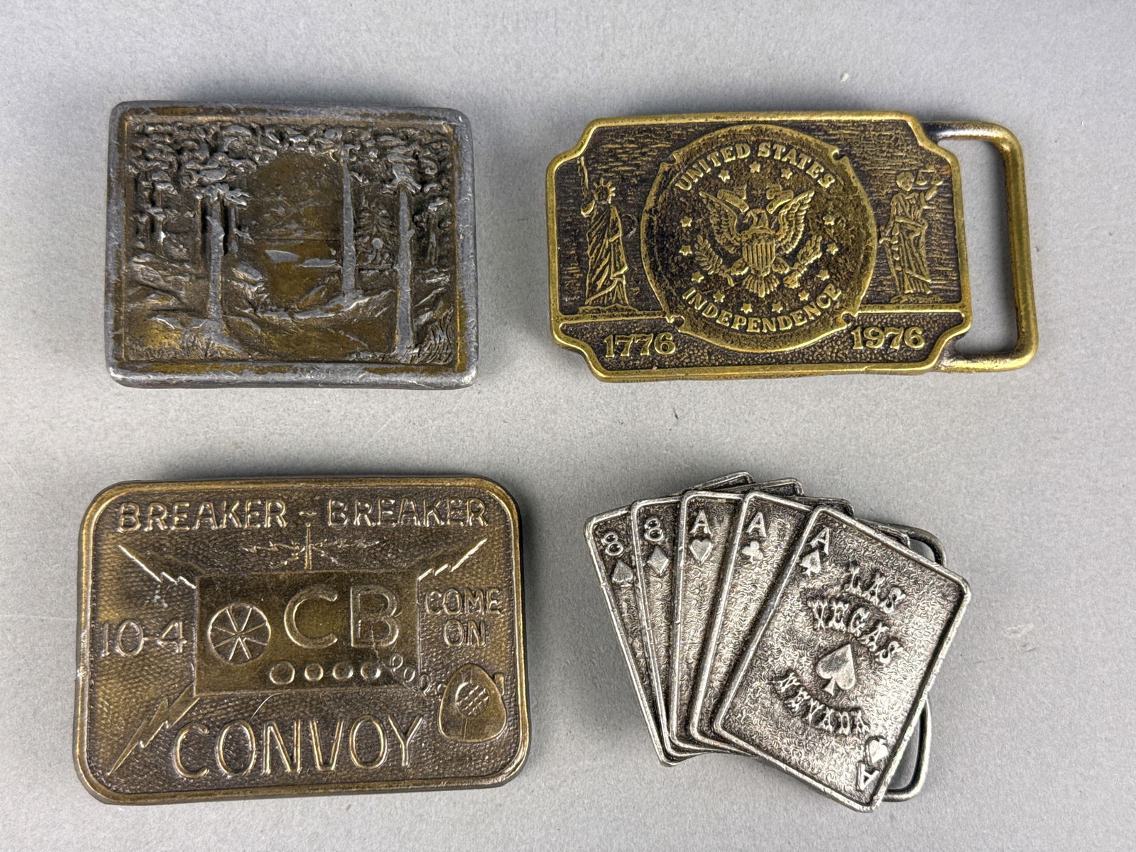 A Collection of Vintage Commemorative and Novelty Belt Buckles including Indiana Metal Craft and: This collection features various cast metal belt buckles from the late 20th century. Included is an Indiana Metal Craft wilderness scene buckle, model A96, depicting a relief-cast forest and lake land
