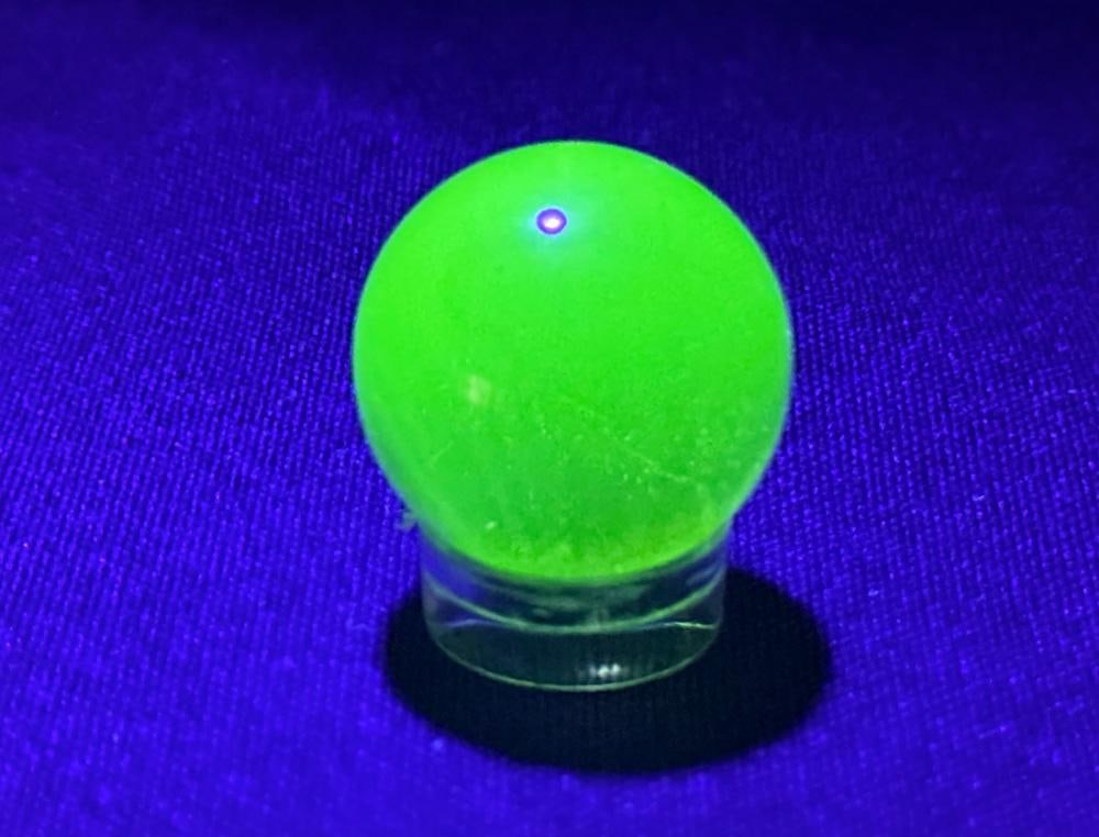 A Vintage Uranium Marble Shooter: The translucent yellowish-green sphere features internal seeds and a distinct yellow swirl or patch. The item exhibits a strong, bright green fluorescence when subjected to ultraviolet light. Diameter