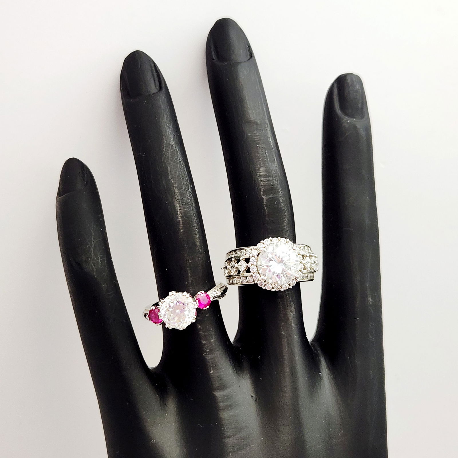 Sterling Cubic Zirconia Rings 9.4 Grams: Two sterling silver rings featuring cubic zirconia and synthetic stones. The first ring is set with a central round clear cubic zirconia flanked by two smaller ruby-colored round stones. The band feat