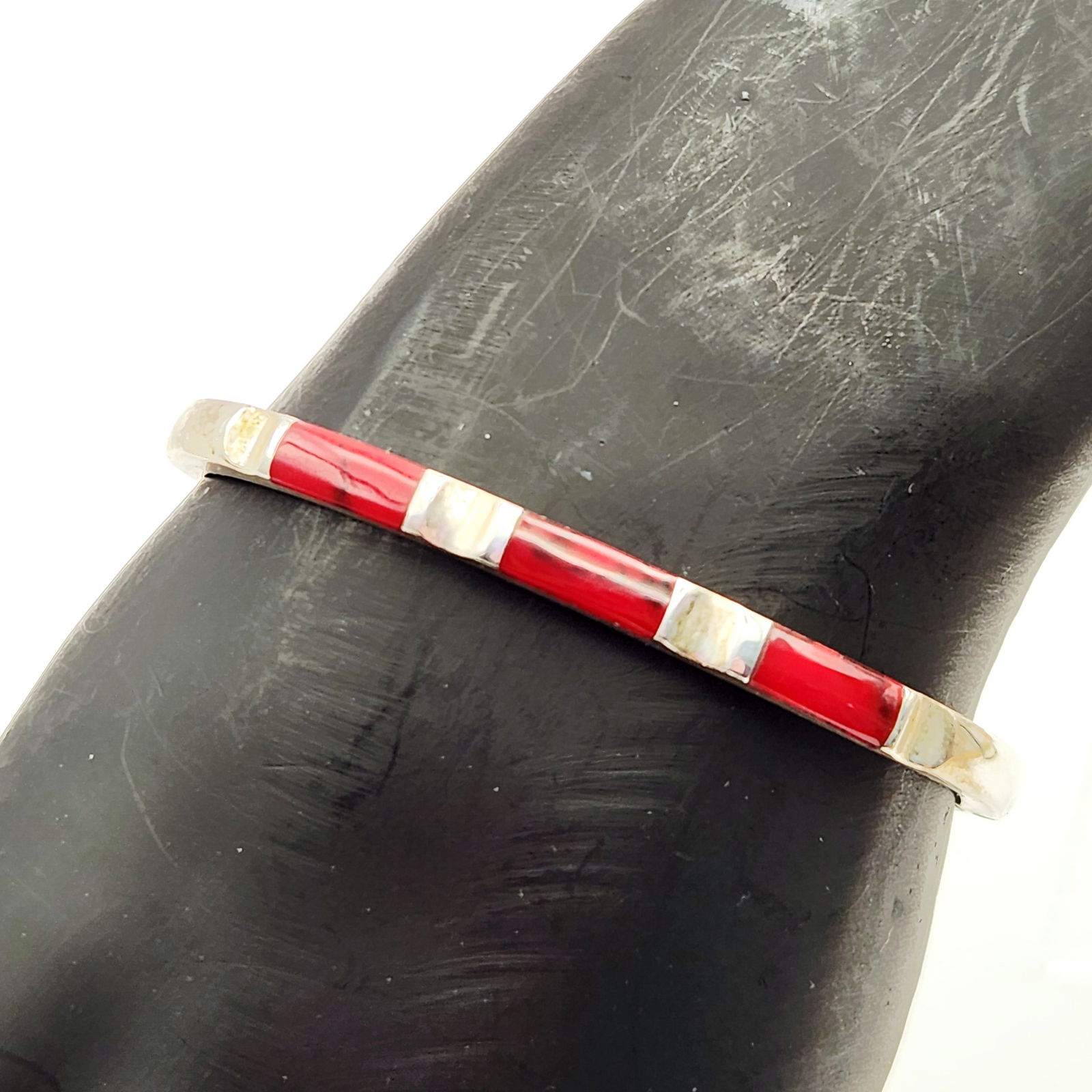 Sterling Coral Inlay Bracelet 19.4 Grams: A Mexican sterling silver hinged bangle bracelet featuring a series of rectangular coral inlays set into a channel. The alternating inlay work consists of a deep red stone. The interior of the band is