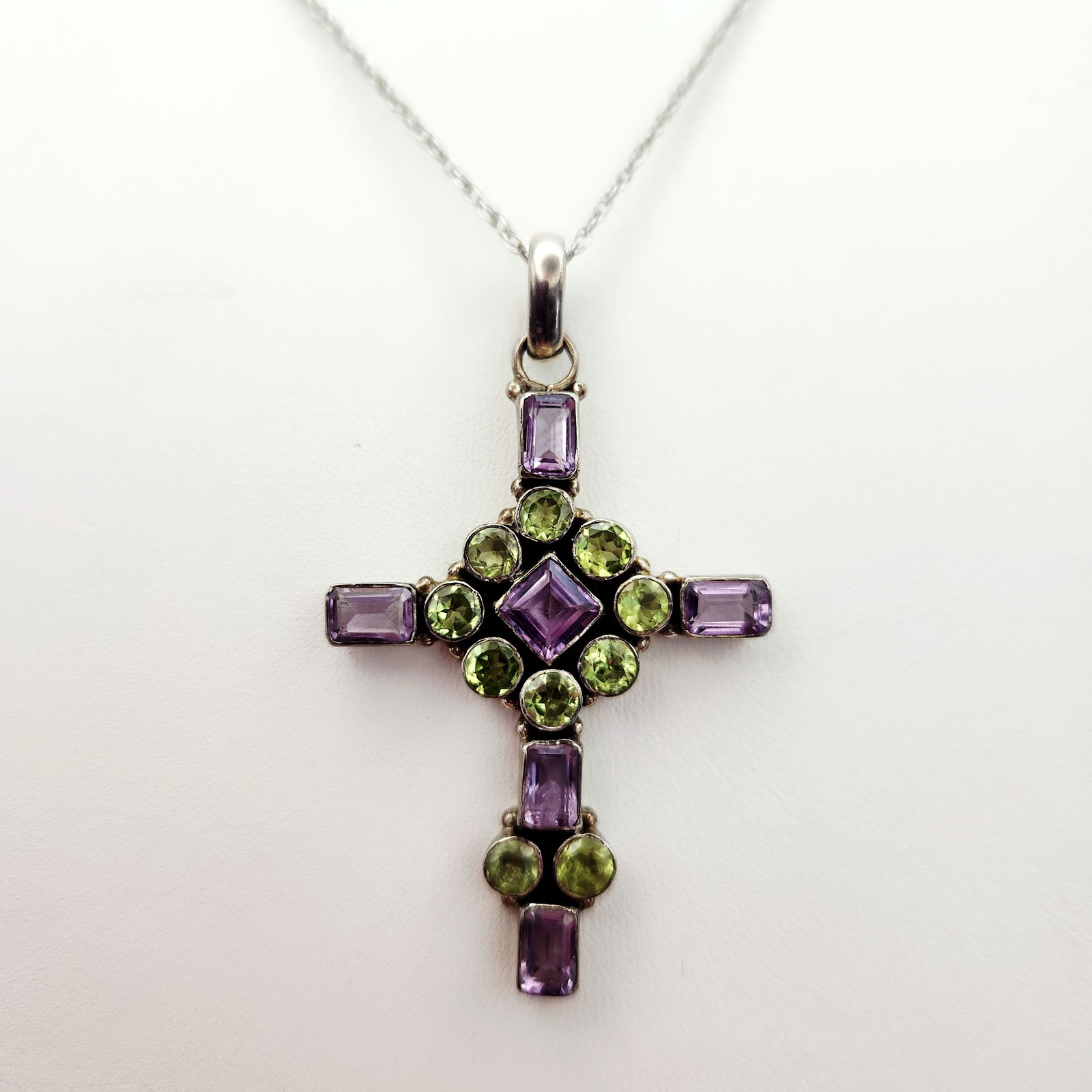 Sterling Amethyst & Peridot Necklace 10.5 Grams: A sterling silver cross pendant featuring faceted purple and green gemstones, including amethyst and peridot. The gemstones are arranged in round, rectangular, and lozenge cuts. The pendant is suspend