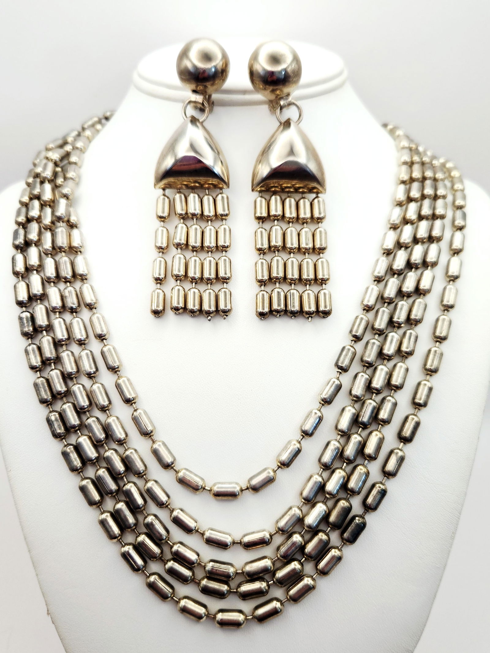 Modernist Silver-Tone Five-Strand Necklace & Coordinating Earrings: This modernist jewelry set consists of a five-strand necklace and a pair of coordinating clip-on dangle earrings, all constructed from polished silver-tone metal. The design features repeated capsule