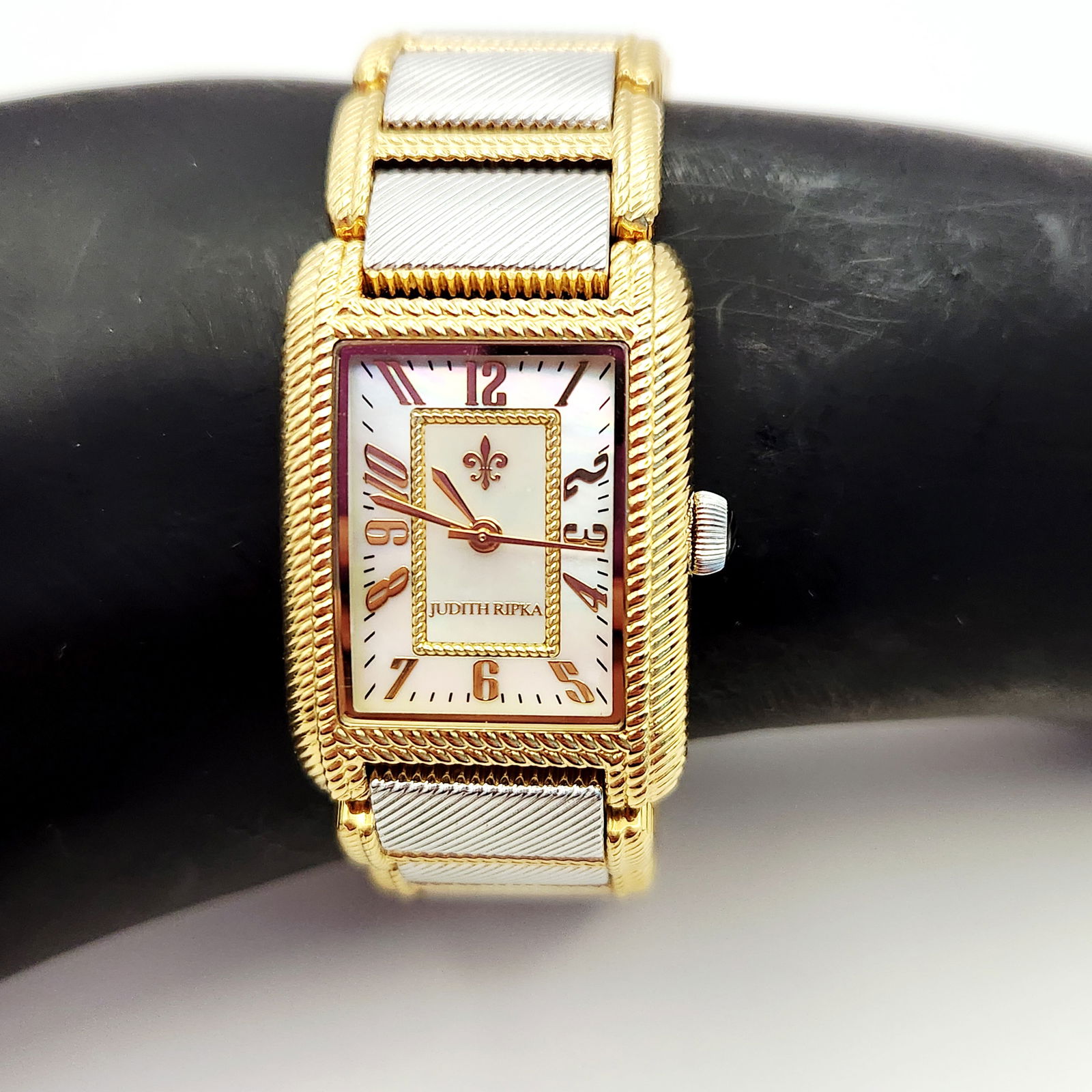 Judith Ripka Ladies Two-Tone Rectangular Wrist Watch: A ladies wrist watch by Judith Ripka featuring a rectangular case with a gold-tone rope-textured bezel. The mother-of-pearl dial displays stylized rose gold-tone Arabic numerals and a fleur-de-lis emb