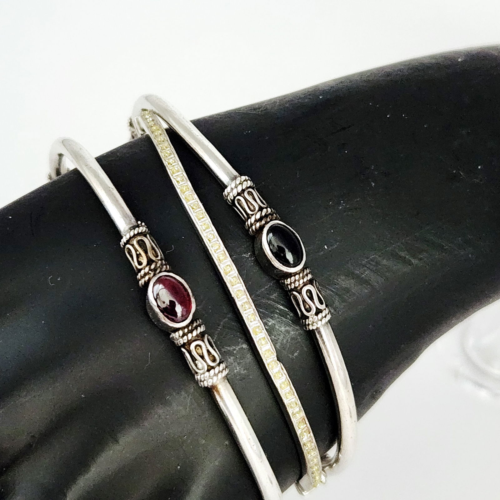 Sterling Silver Gemstone Bangles 25.4 Grams: A collection of three sterling silver bangles. Two bracelets are designed as open cuffs featuring ornate openwork metal details flanking central oval cabochon stones in dark red and black tones. The t