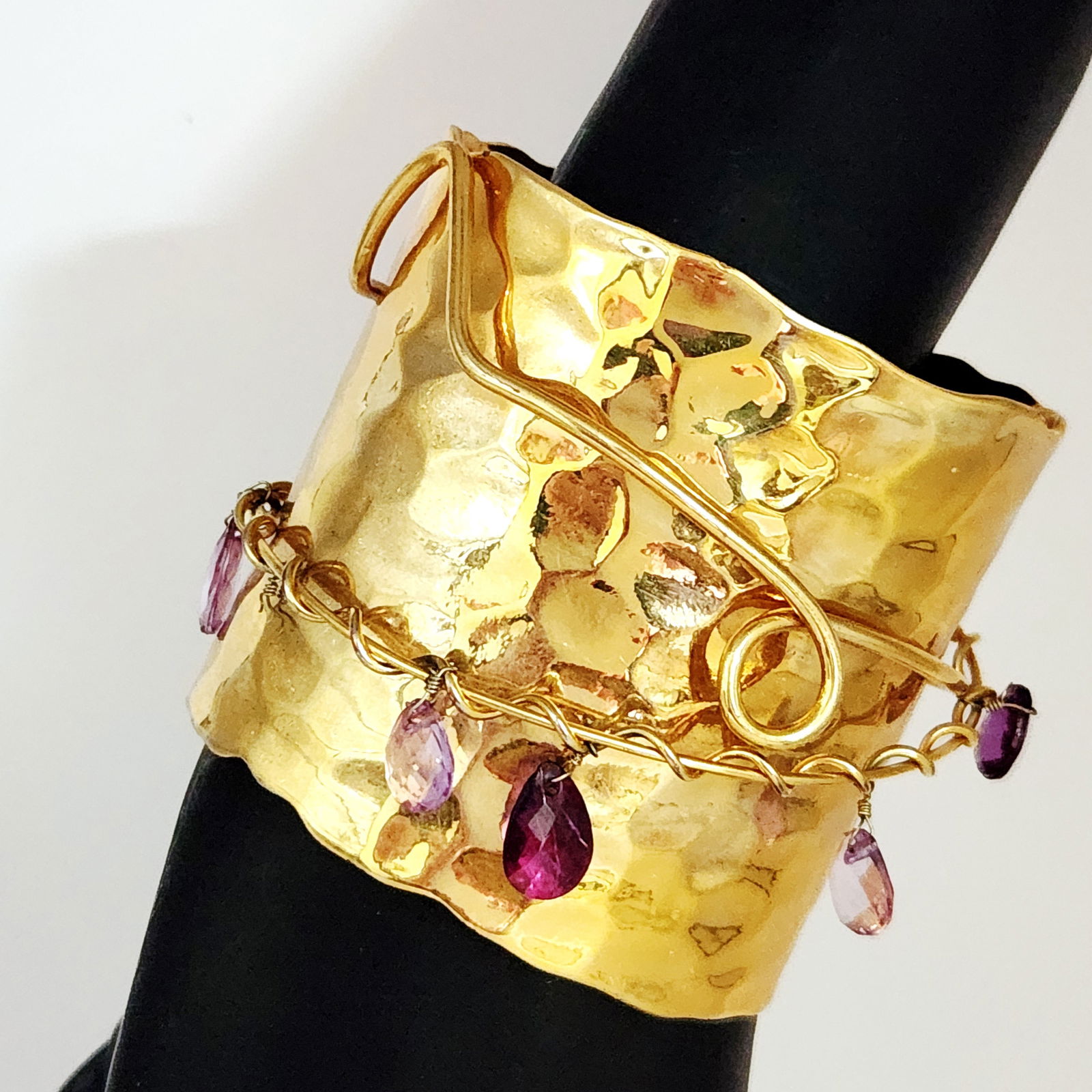 Iman Gold-Tone Hammered Cuff Bracelet with Purple Gemstone Briolettes: A gold-tone cuff bracelet by Iman featuring a wide band with a heavily hammered texture and irregular, wavy edges. The design is accented with decorative wire-wrapped overlays and a series of dangling
