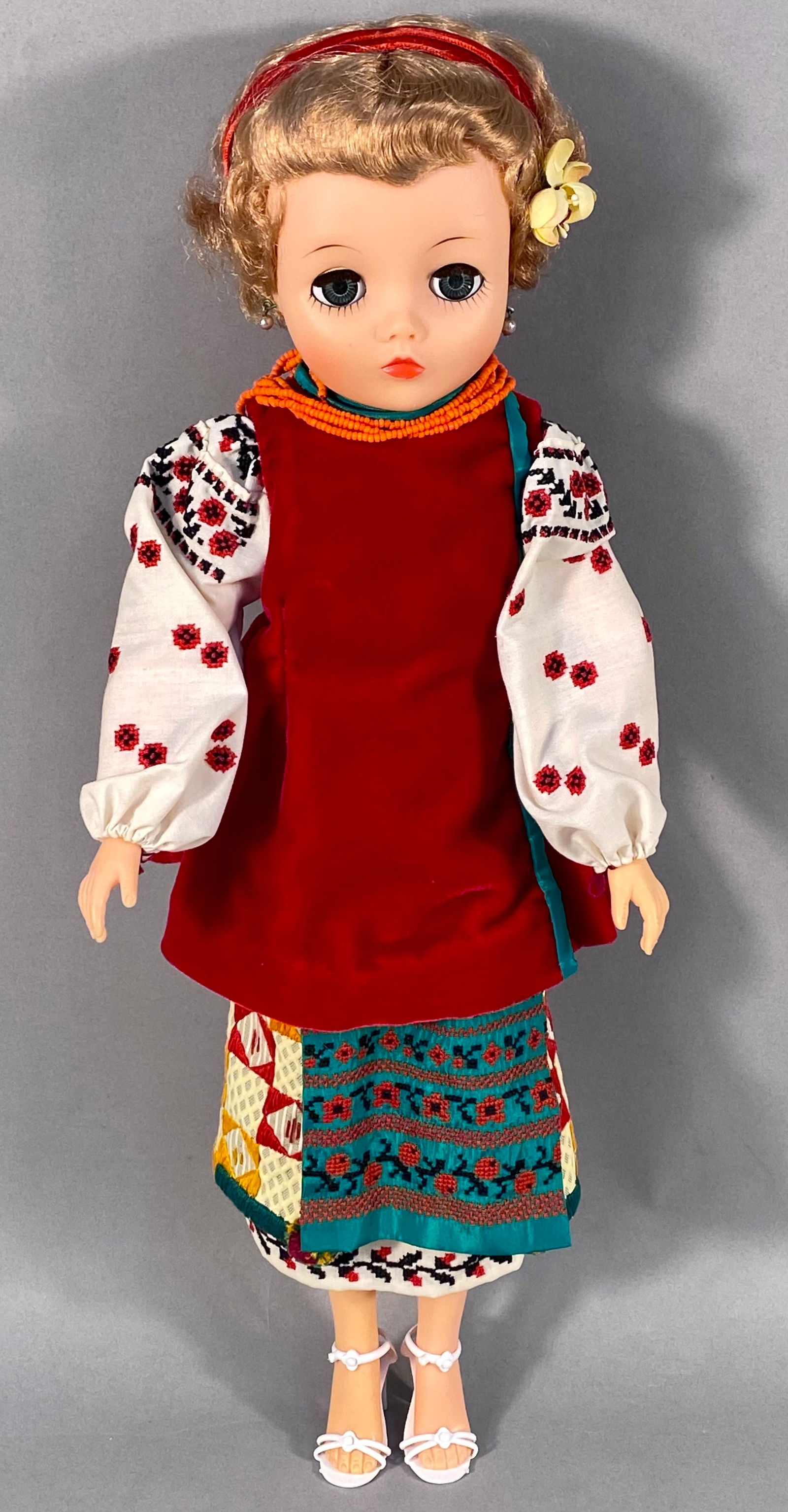 Vintage Uneeda Ukrainian Doll in Traditional Folk Costume: This vintage vinyl doll, manufactured by Uneeda, is dressed in a traditional Ukrainian folk costume. The doll features a vinyl head with rooted blonde hair styled with a red headband and a yellow fabr