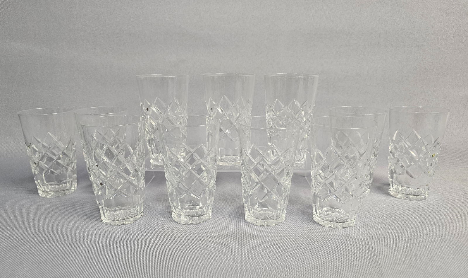 A Collection of 11 20th-Century Cut Crystal Juice Tumblers: A collection of clear cut-glass highball tumblers. Each vessel is constructed of crystal with a tapered, cylindrical form. The exterior body features a prominent diamond-cut pattern, while the base