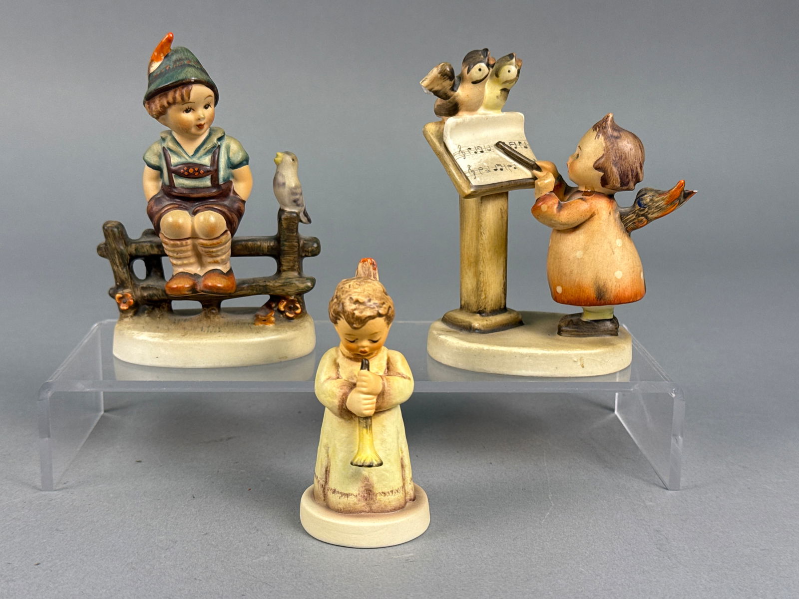 A Trio of Goebel M.I. Hummel Hand-Painted Porcelain Figurines Including Bird Duet: A trio of Goebel M.I. Hummel hand-painted porcelain figurines, including the Bird Duet pattern. The collection features a boy in traditional Bavarian dress seated on a fence with a bird marked 1938, a