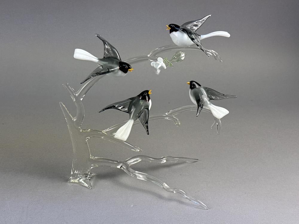 A Group of Murano Glass Birds on a Branch: This Murano glass sculpture features a group of birds perched on a clear glass branching base. The hand-blown work includes birds with black, white, and gray bodies and yellow beaks, alongside a singl