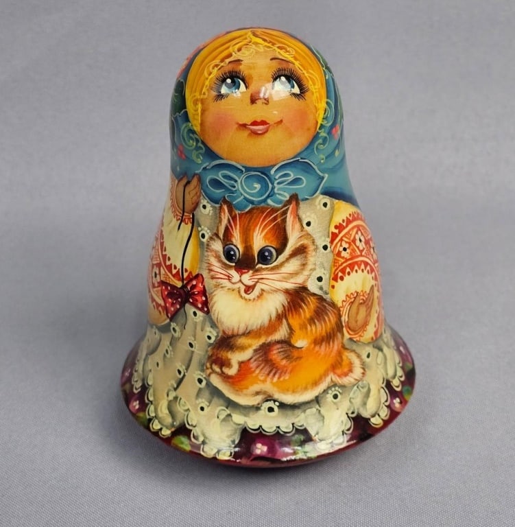 A Russian Matryoshka Style Hand-Painted Wooden Bell: A hand-painted wooden decorative bell executed in a traditional Russian Matryoshka folk art style. The object features a glossy lacquered finish and depicts a young blonde girl in a blue headscarf hol