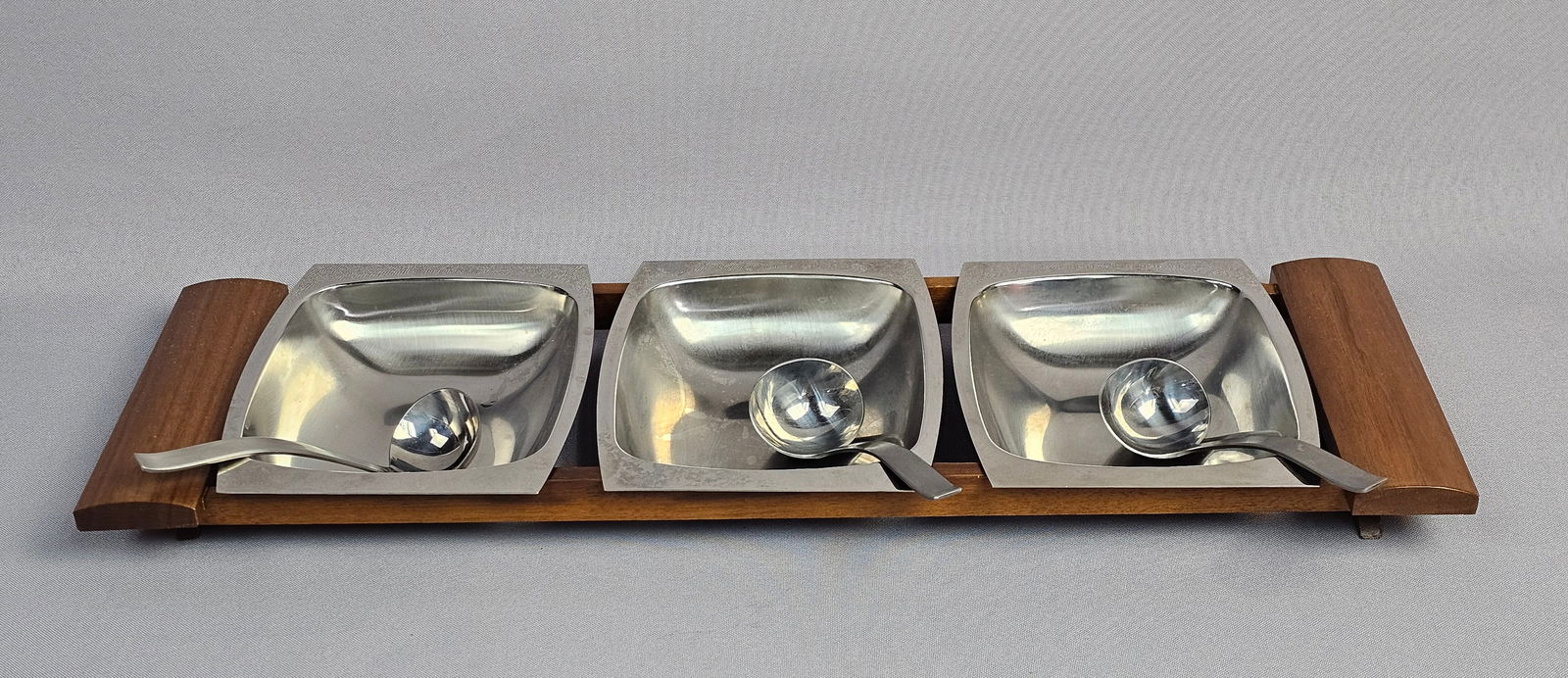 A Mid-Century Modern Stainless Steel and Teak Serving Set: A mid-century modern serving set comprising stainless steel square-form bowls and matching ladles. The metal components are fitted within a linear teakwood rack designed with raised, curved end handle