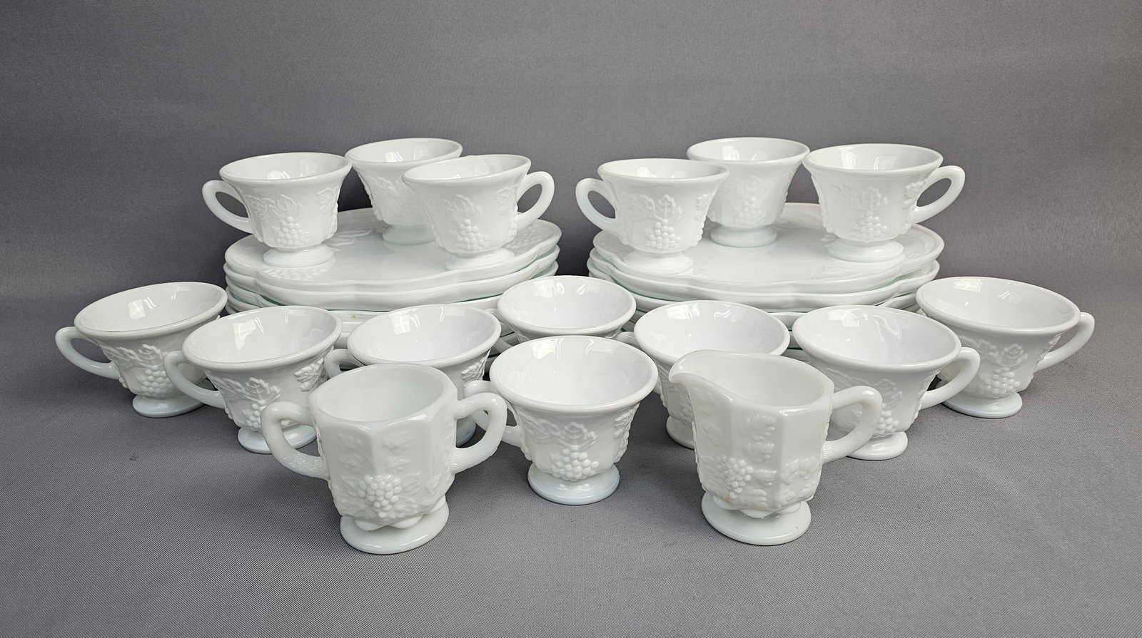 A Collection of Indiana Glass Colony Harvest Milk Glass Snack Set Items: This collection of white milk glass items is produced in the Colony Harvest pattern by Indiana Glass, featuring embossed grape clusters and vine leaves. The set includes several scalloped-edge luncheo