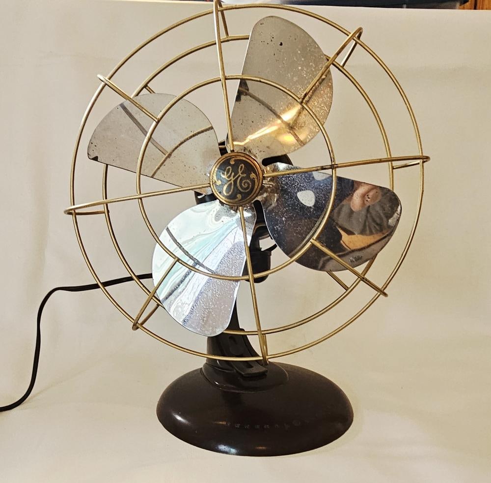 A General Electric 12 Inch Stationary Brass Bladed Desk Fan: A General Electric stationary electric desk fan, circa 1920. The fan features a dark brown motor housing and a matching circular base embossed with the General Electric name and monogram. It is equipp