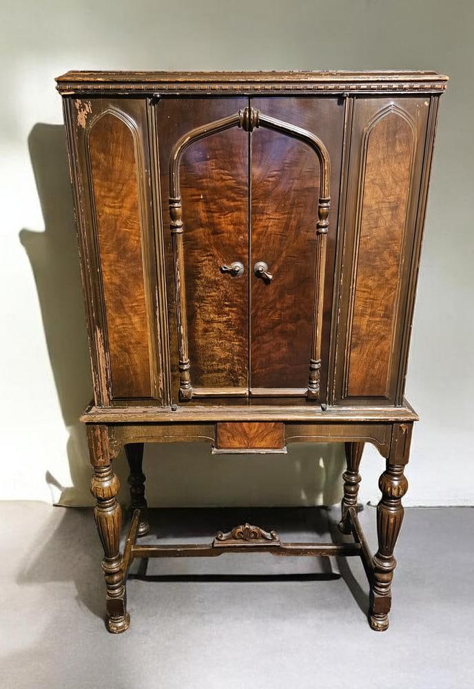 Antique Radio Cabinet by Adler-Royal: The cabinet features Tudor and Gothic Revival design elements, including pointed arch panels and decorative moldings. The structure is supported by four turned and fluted legs connected by a stretcher