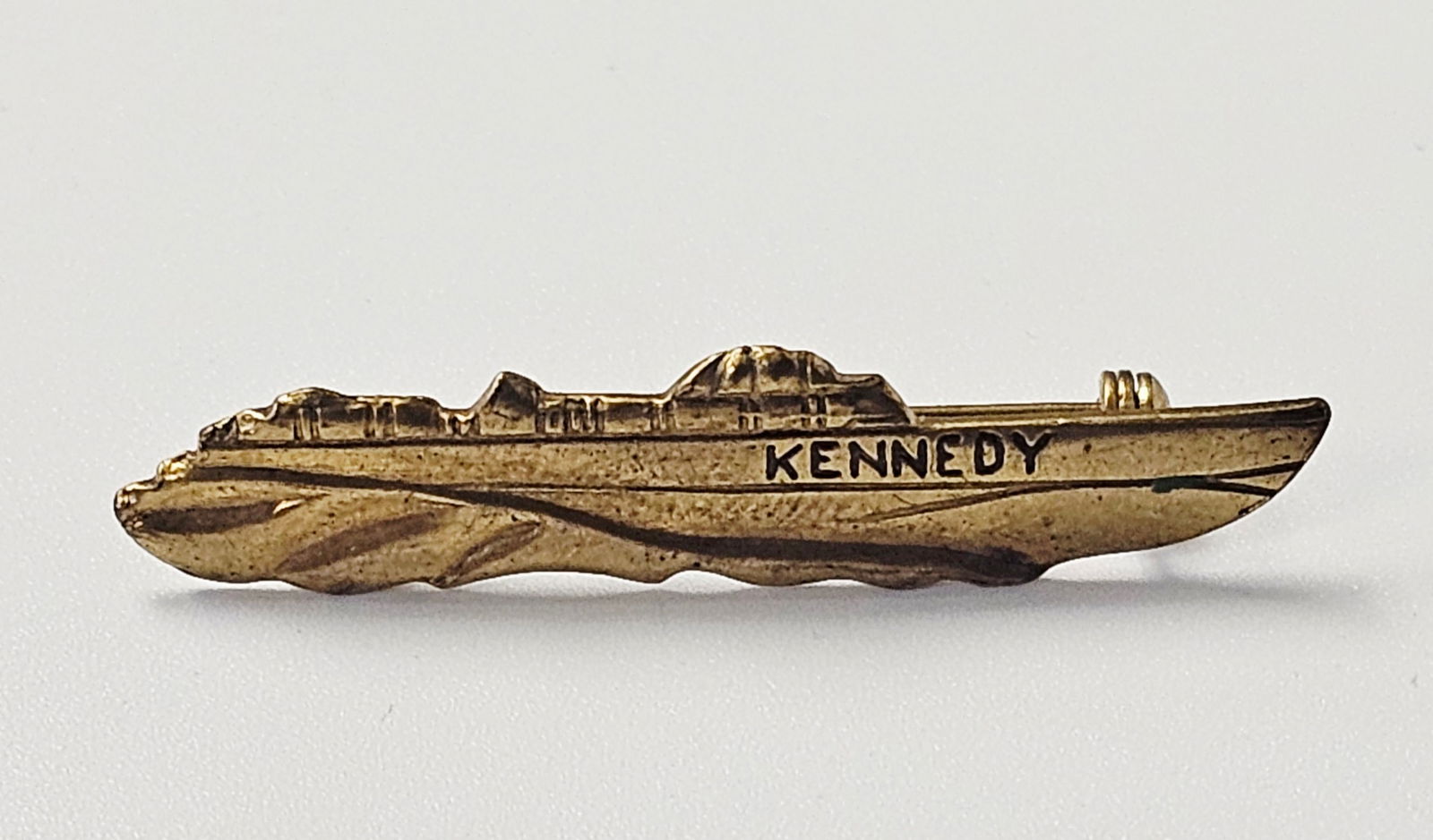 John F. Kennedy 1960 PT-109 Presidential Campaign Pin: A John F. Kennedy PT-109 presidential campaign pin, cast in gold-toned textured metal. The piece is modeled in the form of a naval patrol torpedo boat, featuring the name KENNEDY prominently stamped