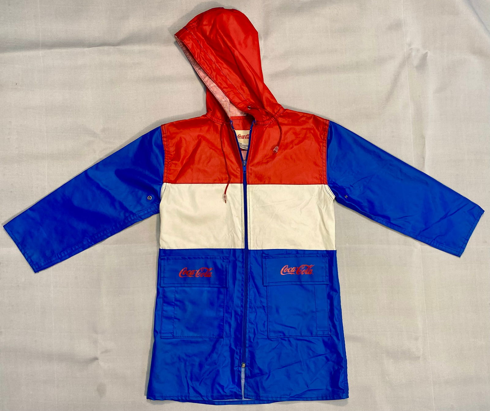 Coca-Cola Child's Rubberized Hooded Raincoat, Color-Block Design, c. 1970s-1980s: This vintage promotional garment is a child's hooded raincoat featuring a vibrant red, white, and blue horizontal color-block motif. The piece is constructed from a durable PVC exterior with a soft po