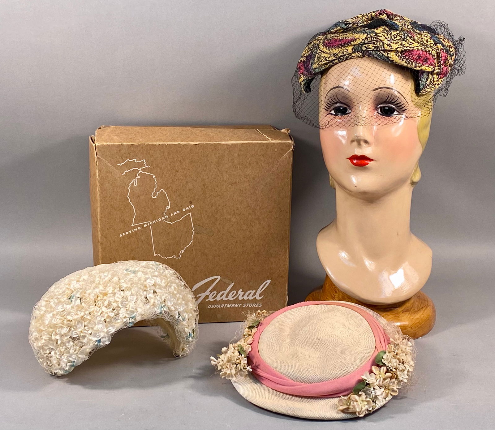 A Group of Mid-20th Century Women's Hats and Accessories including a Federal Department Stores Box: A group of mid-20th-century women's millinery accessories. The collection includes a paisley-patterned fabric turban featuring a black net veil. Also included is a white pillbox hat with a floral-text