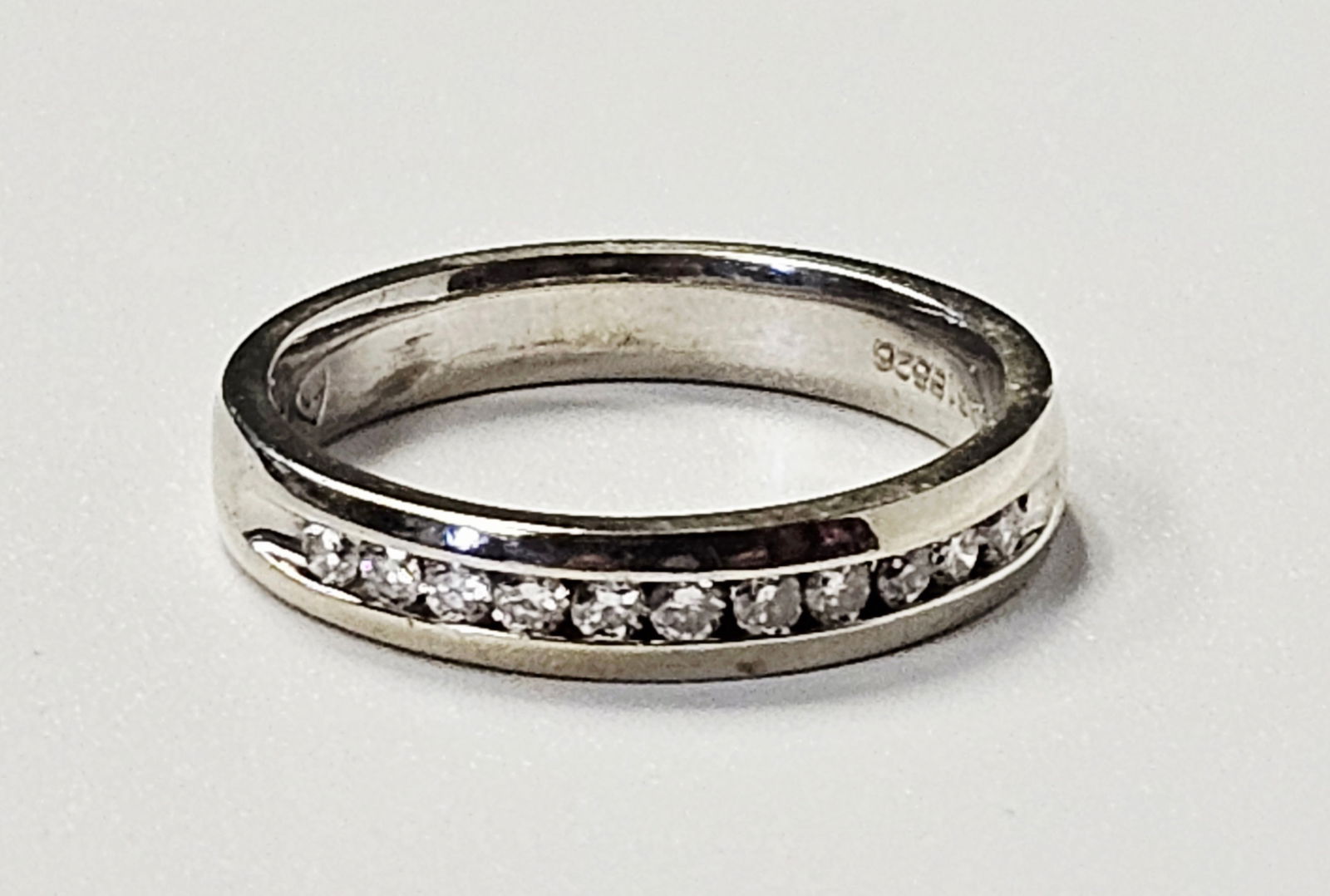 18k White Gold & Diamond Channel-Set Band 4.4 Grams: This ring is crafted in 18k white gold and features a channel-set row of round-cut colorless stones that appear to be diamonds. The interior of the shank is hallmarked 18K and includes a maker's mark