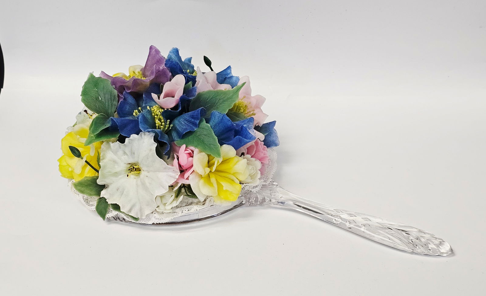 Vintage Lucite Hand Mirror Floral Arrangement: This handheld mirror features a clear, molded acrylic handle with stylized leaf and geometric diamond patterns. The reverse side is decorated with a three-dimensional floral arrangement composed of fa