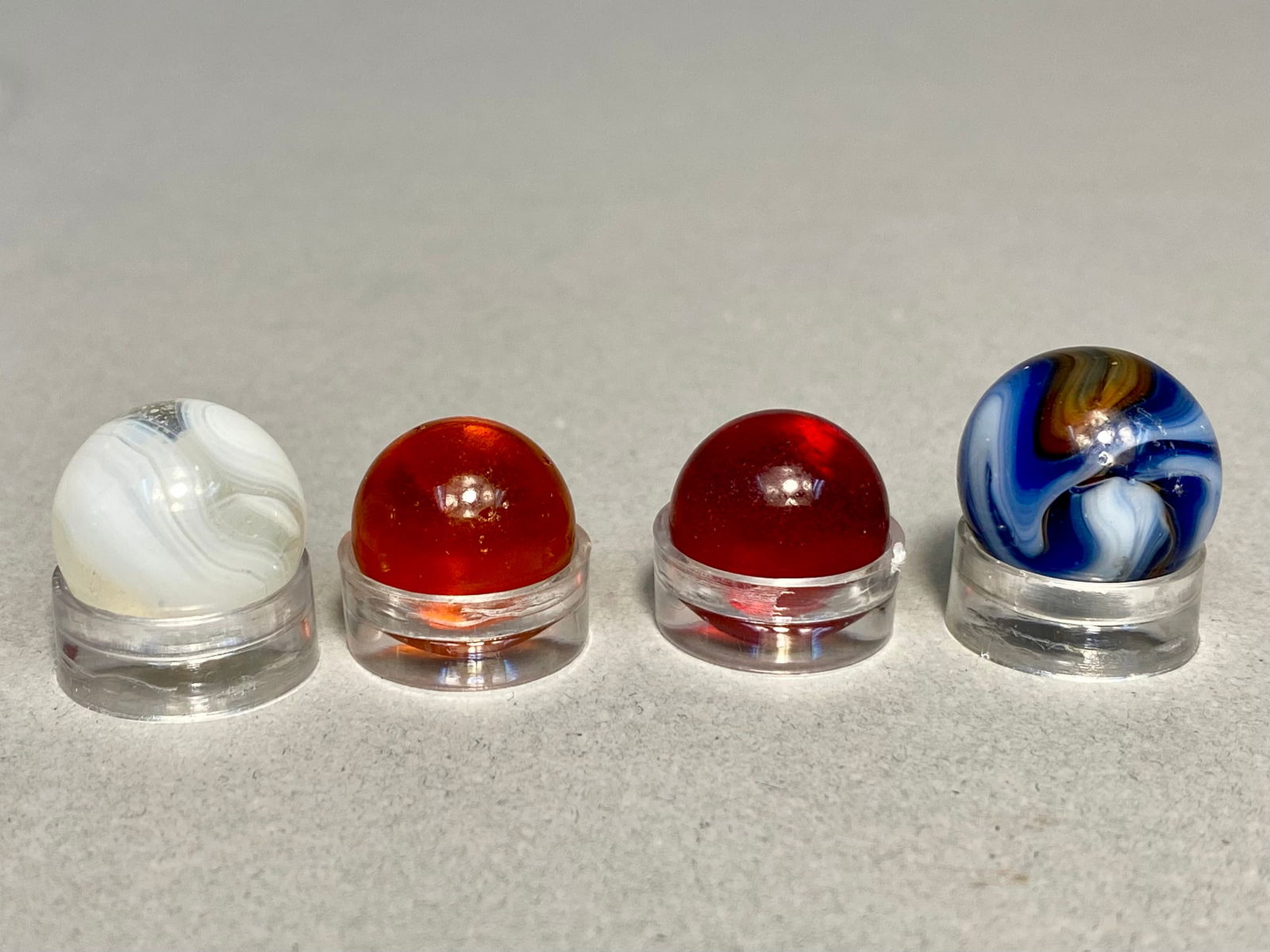 Collection of Four Vintage Machine-Made Glass Marbles, Assorted Styles, mid-20th Century: This lot consists of four vintage machine-made glass marbles representing various popular mid-century styles. The collection includes two translucent "clearies" in ruby red and amber tones,