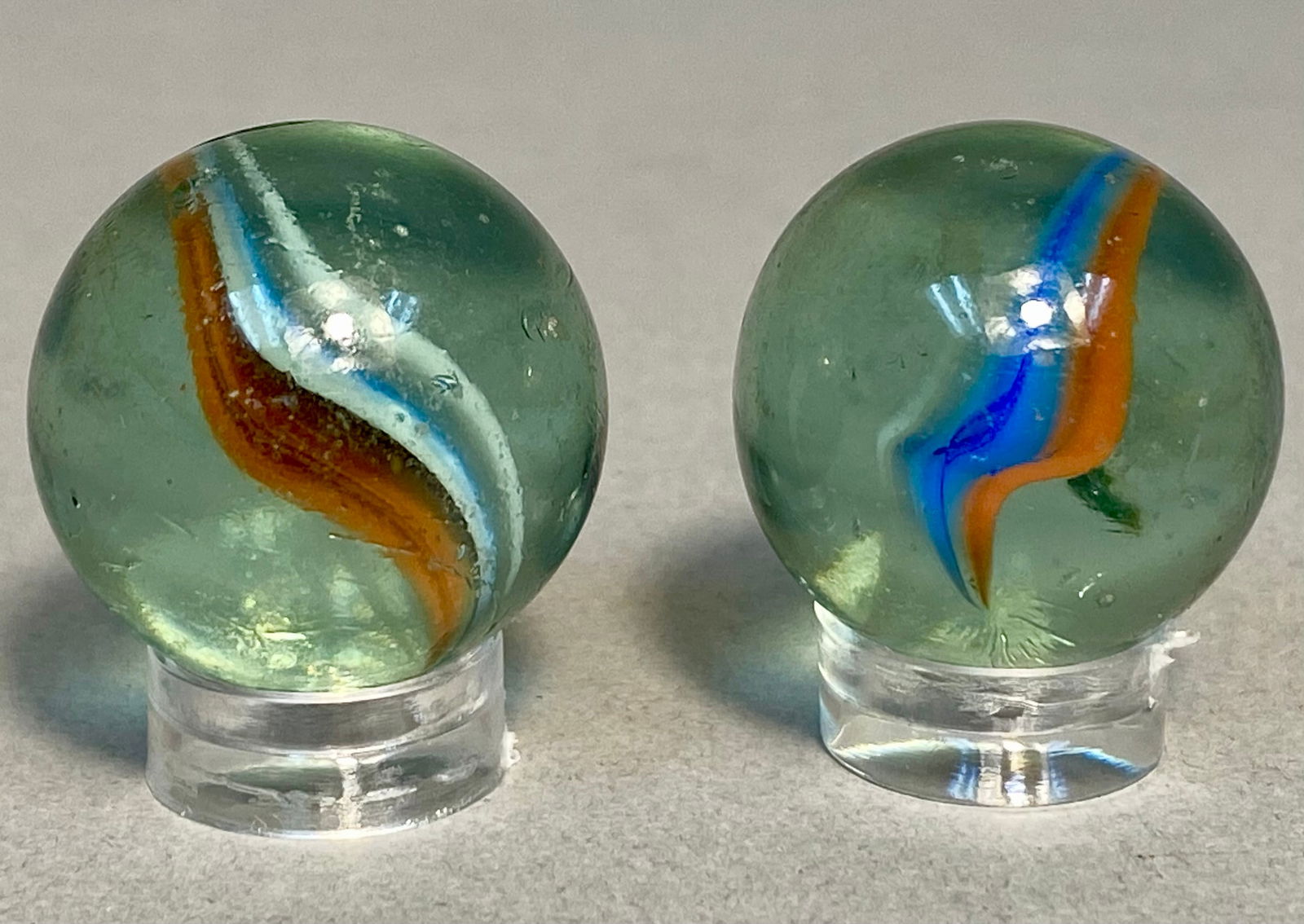 A Pair of Vintage Cat's Eye Glass Marbles with Blue and Orange Ribbons, mid-20th Century: This lot features two vintage cat's eye style glass marbles, characteristic of mid-20th-century toy production. Each marble is composed of a clear, pale aqua-green glass base housing internal multi-va