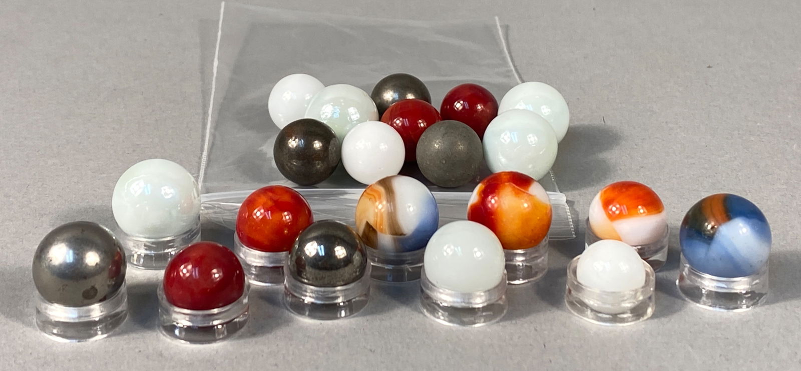 Collection of Vintage Glass and Metallic Marbles, Various Swirl and Solid Patterns, mid-20th Century: A diverse assortment of vintage marbles featuring a variety of materials and designs. The collection includes machine-made glass marbles in solid white, solid red, and translucent pale green, alongsid