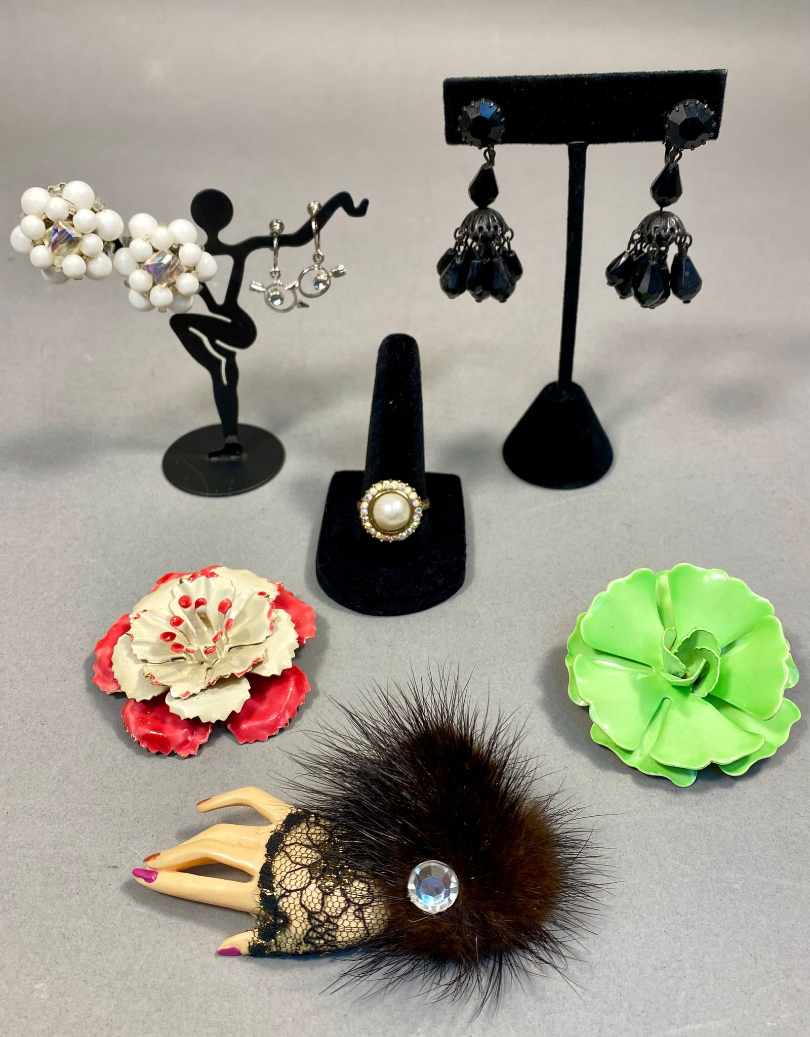 An Assortment of Mid-Century Costume Jewelry Including a Novelty Hand Brooch: An assortment of mid-20th-century costume jewelry. The collection includes a novelty hand-shaped brooch featuring painted fingernails, a black lace cuff, brown faux fur, and a central colorless rhines