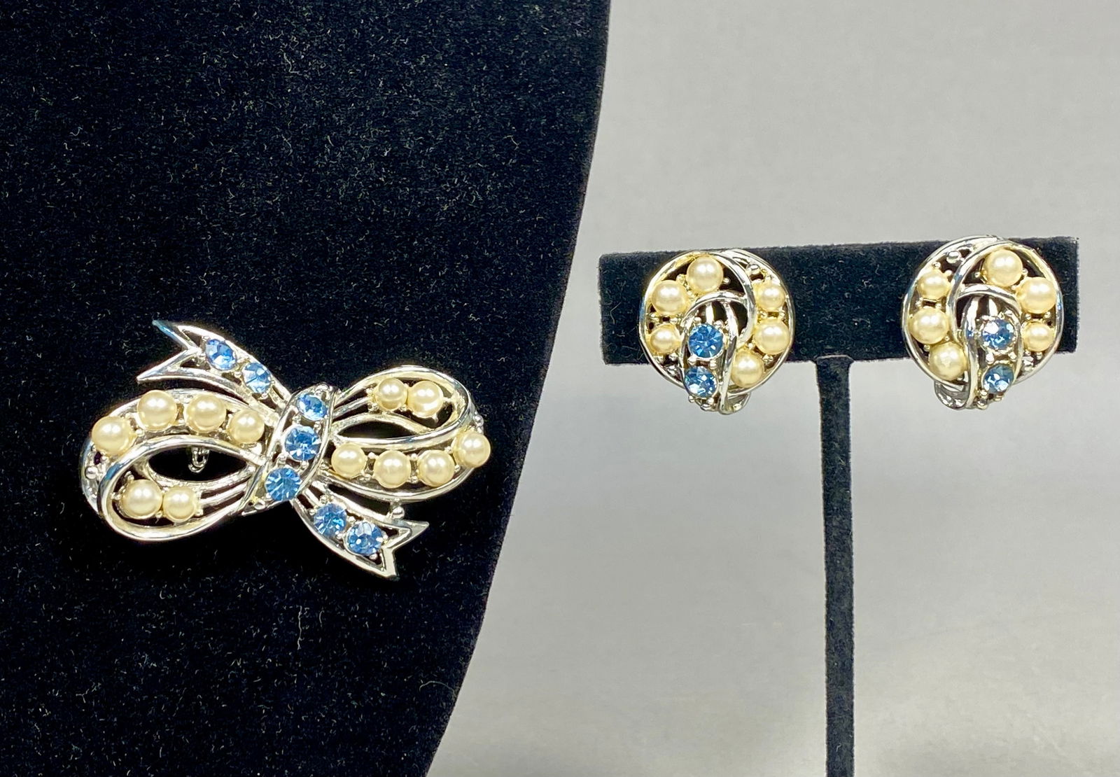 Silver-Tone Bow Brooch and Earring Set with Blue Rhinestones and Simulated Pearls by Coro: This vintage costume jewelry set includes a stylized bow-shaped brooch and a coordinating pair of circular earrings. The pieces are constructed from a silver-tone base metal. The brooch features a mul
