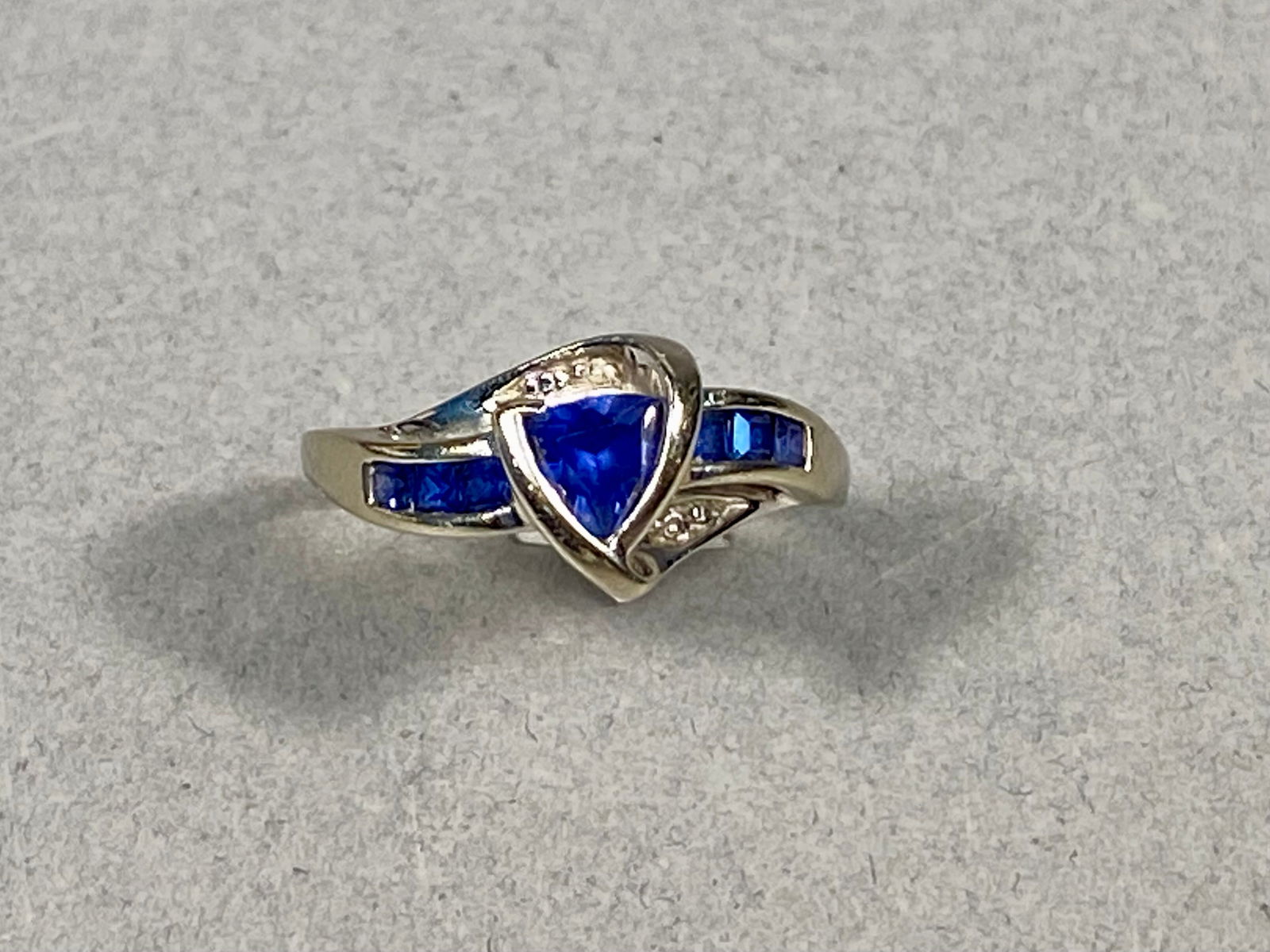 10 Karat Gold Bypass Ring with Trillion Cut Blue Gemstone and Accents, 2.13g: This bypass-style ring is crafted in 10 karat gold and features a central trillion-cut blue gemstone that appears to be a sapphire. The design is further accented by six channel-set square-cut blue ge