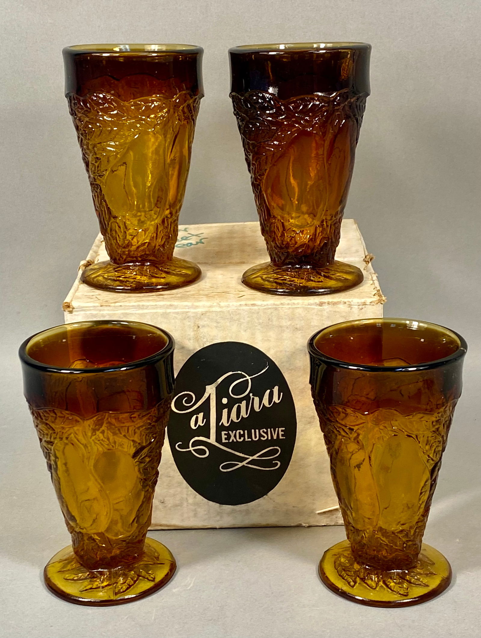 Tiara Exclusive, Pressed Amber Glass, Footed Tumblers: A set of four vintage footed tumblers produced for the Tiara Exclusive line by Indiana Glass Company. These pieces are executed in a warm amber glass, often marketed as Antique Gold. The design featur