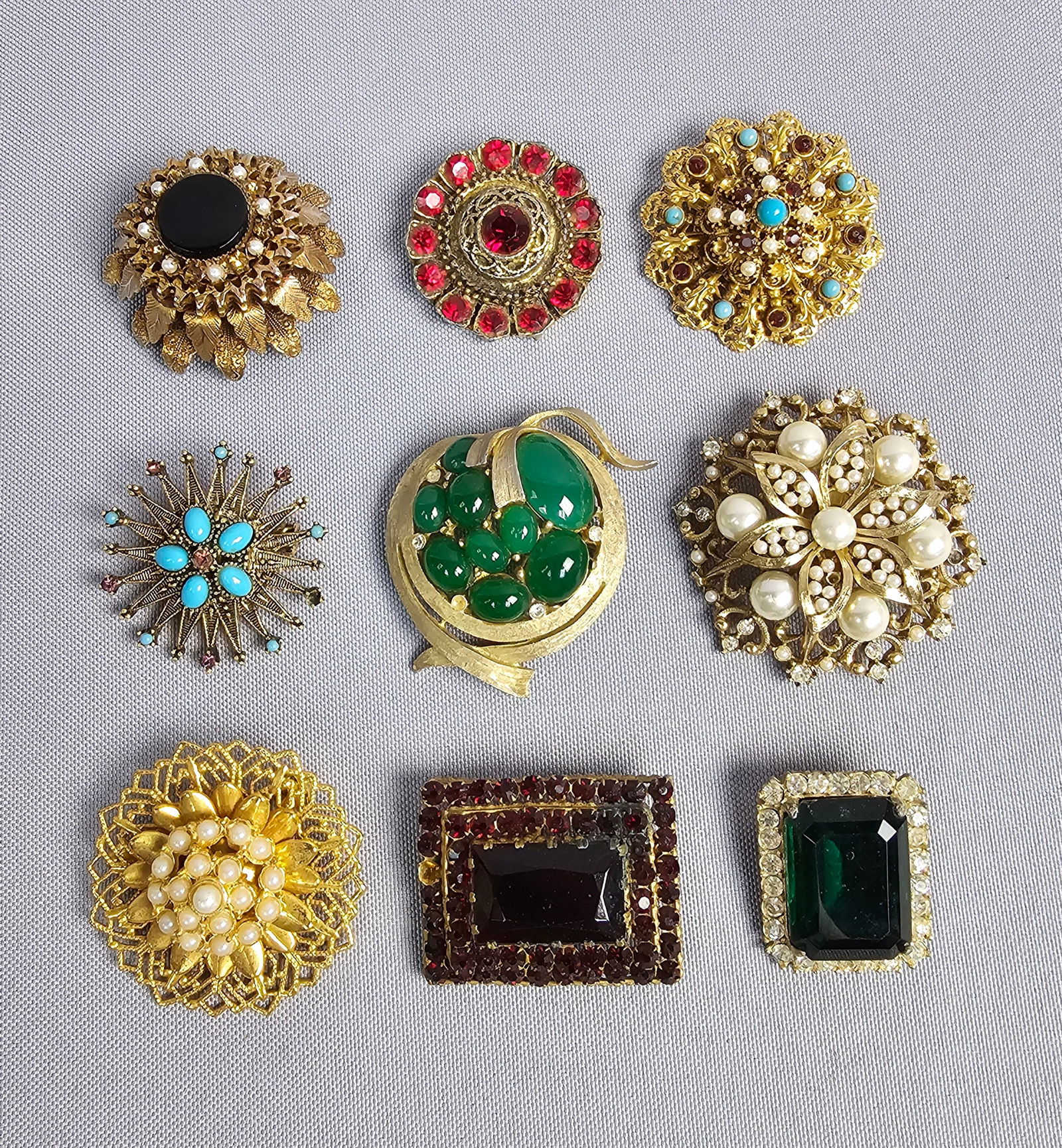 Gold-Tone Floral and Geometric Costume Brooches: A group of nine vintage costume jewelry brooches featuring floral, geometric, and modernist motifs. Featured is a tiered rectangular brooch with a large emerald-cut red glass stone and red rhinestone