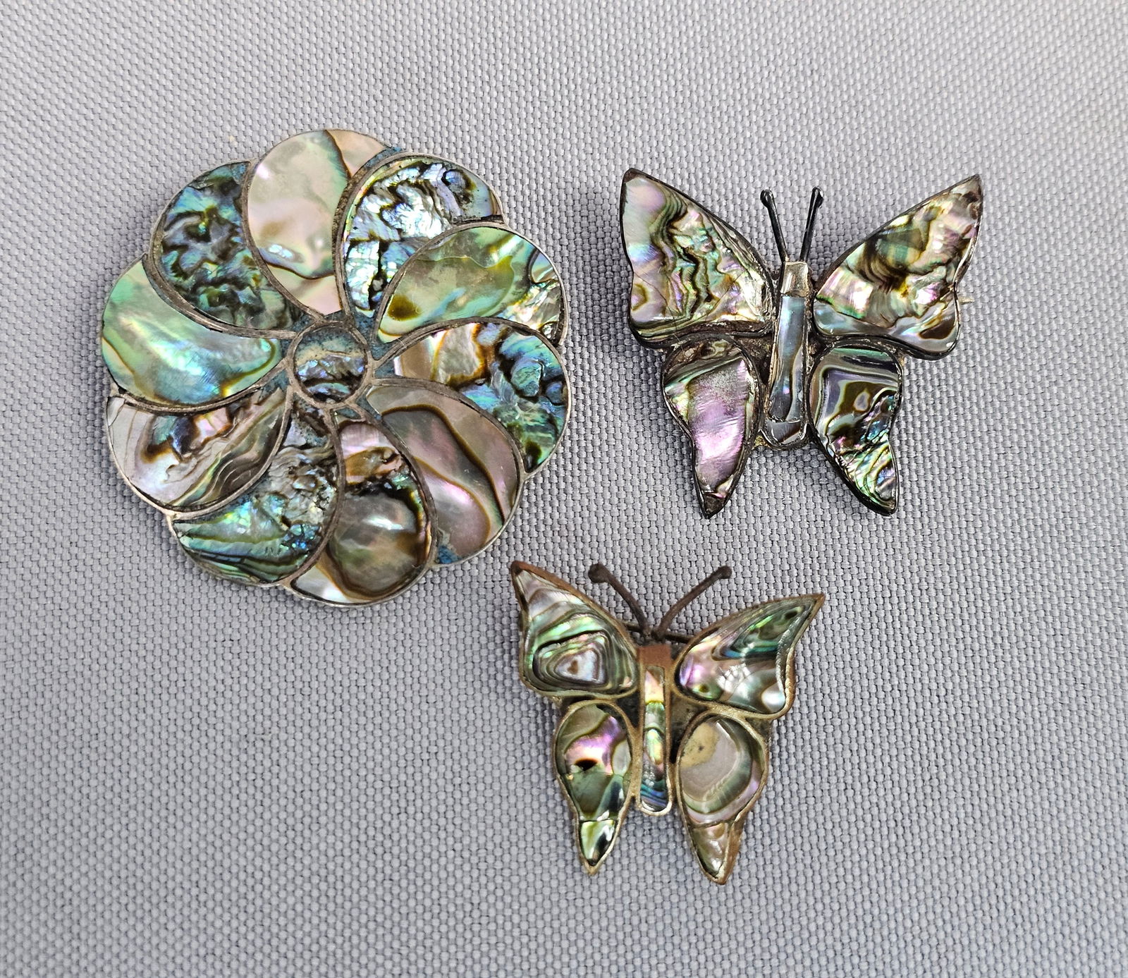 Three Sterling Silver and Abalone Inlay Pinwheel and Butterfly Brooches: This lot features three vintage Mexican jewelry pieces showcasing traditional Taxco craftsmanship. The first is a circular pinwheel or floral swirl brooch that also functions as a pendant, featuring r