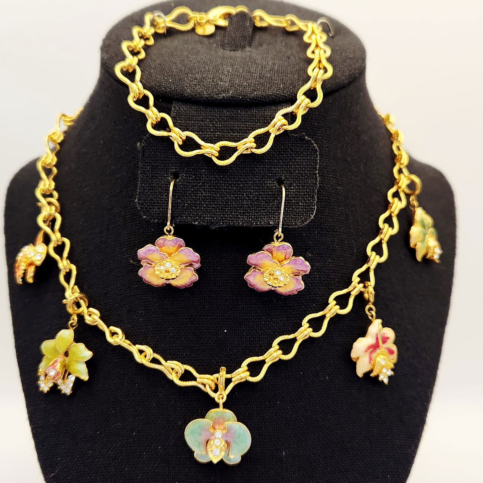 Nolan Miller Orchid Enamel Jewelry Set: A trio of gold-tone jewelry by Nolan Miller featuring colorful enamel orchid charms with rhinestone accents, including a 20-inch necklace, a 7.5-inch bracelet, and a pair of earrings. In good vintage
