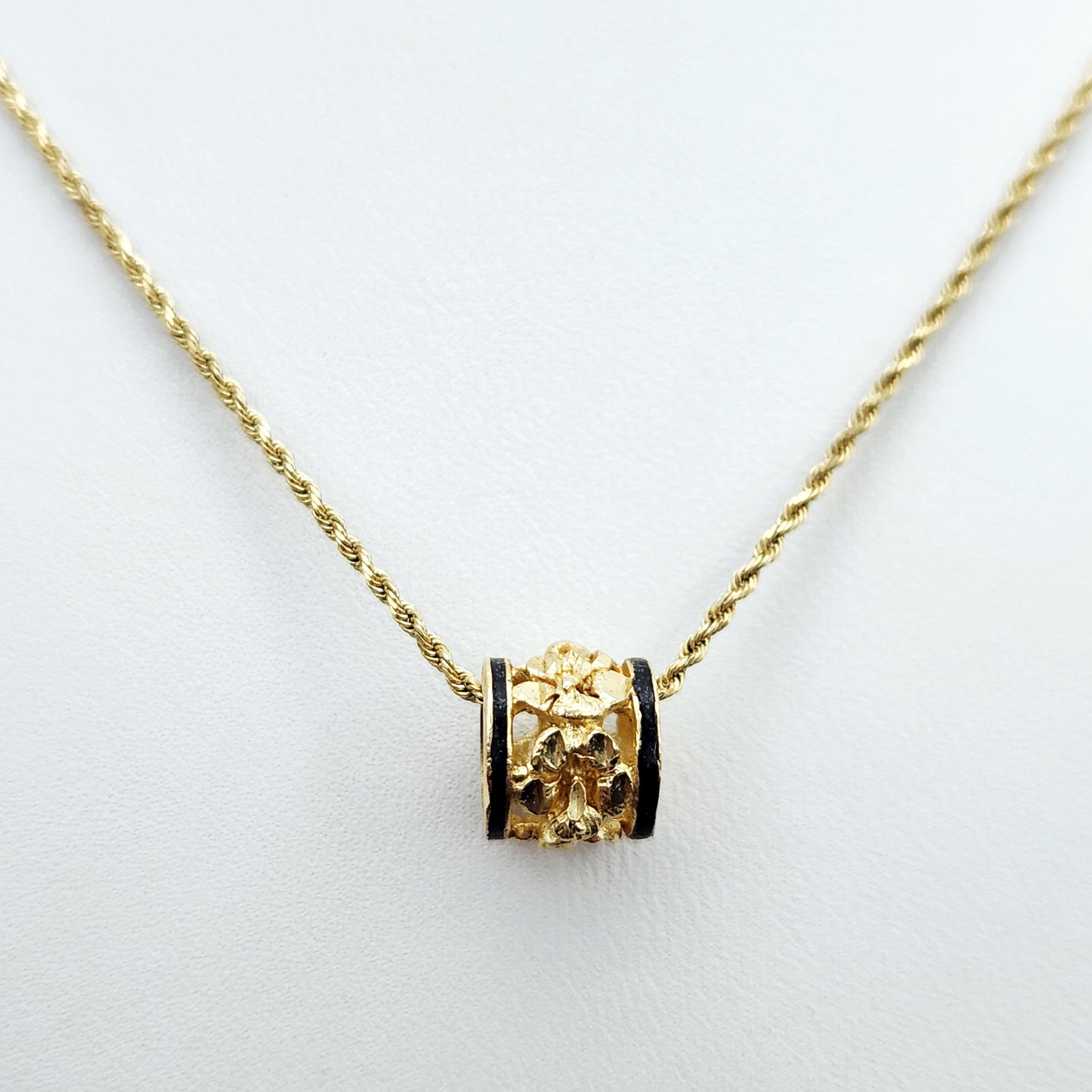 14K Gold & Black Enamel Necklace 5.1 Grams: This necklace is crafted in 14K yellow gold and features a barrel-style slide pendant suspended from a fine rope-link chain. The pendant is designed with a central openwork band of textured gold in a