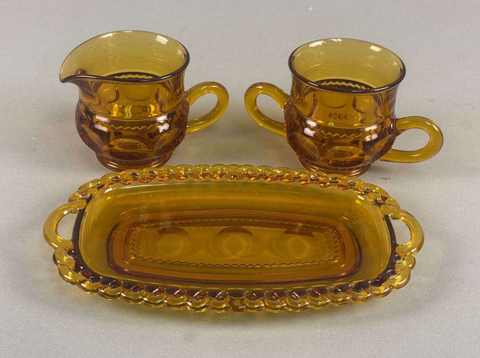 A Trio of Amber Pressed Glass Sugar and Creamer Set Pieces with Tray: Inidana Glass Attributed to the Kings Crown pattern, this trio of amber pressed glass includes a single-handled creamer, a double-handled open sugar bowl, and an oval serving tray. The set is crafted
