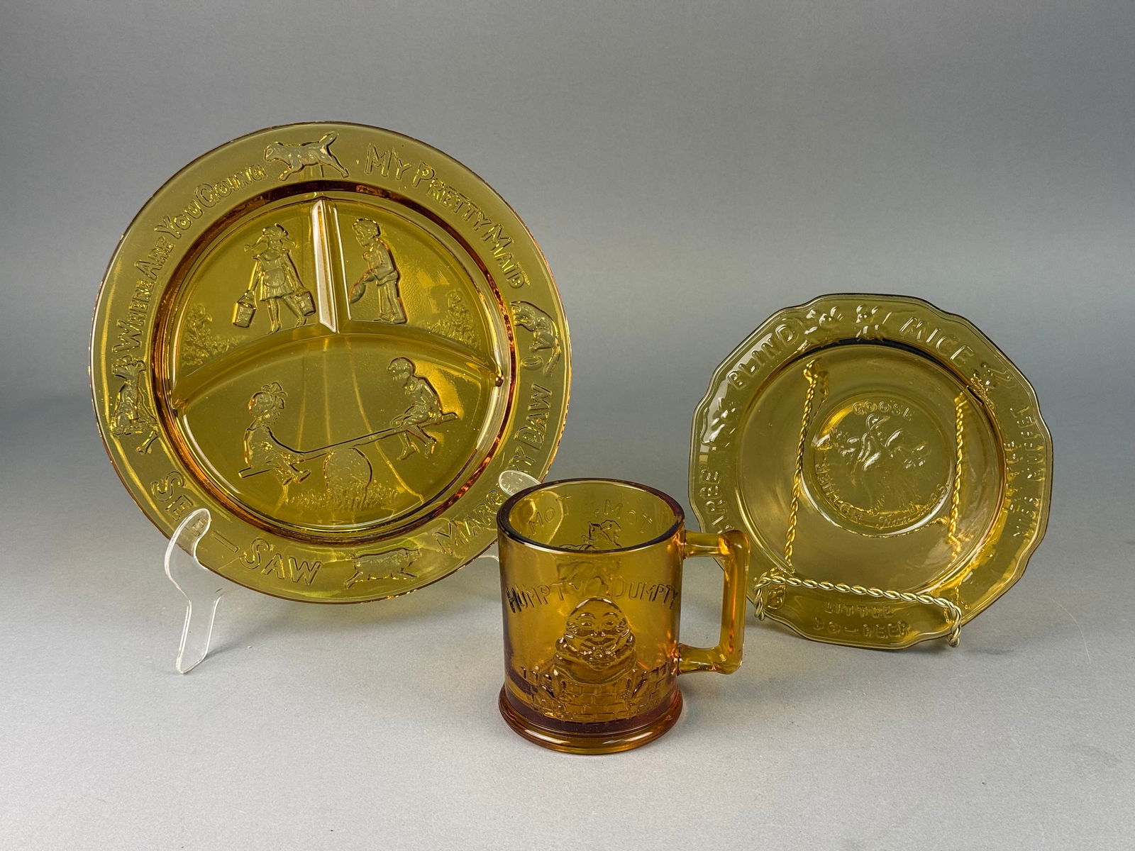 A Trio of Tiara Exclusives Amber Glass Nursery Rhyme Child Tableware: A trio of amber pressed glass child's tableware pieces produced for Tiara Exclusives. The collection features various embossed nursery rhyme motifs with integrated text. The set includes a three-part