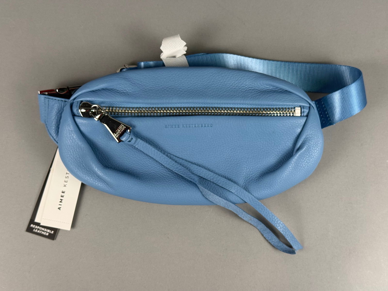 Aimee Kestenberg Milan Pebbled Leather Belt Bag in Cornflower Blue: This Aimee Kestenberg Milan belt bag is crafted from cornflower blue pebbled leather with a tonal nylon webbing strap. The bag features a main zippered compartment with an extra-long leather tassel pu