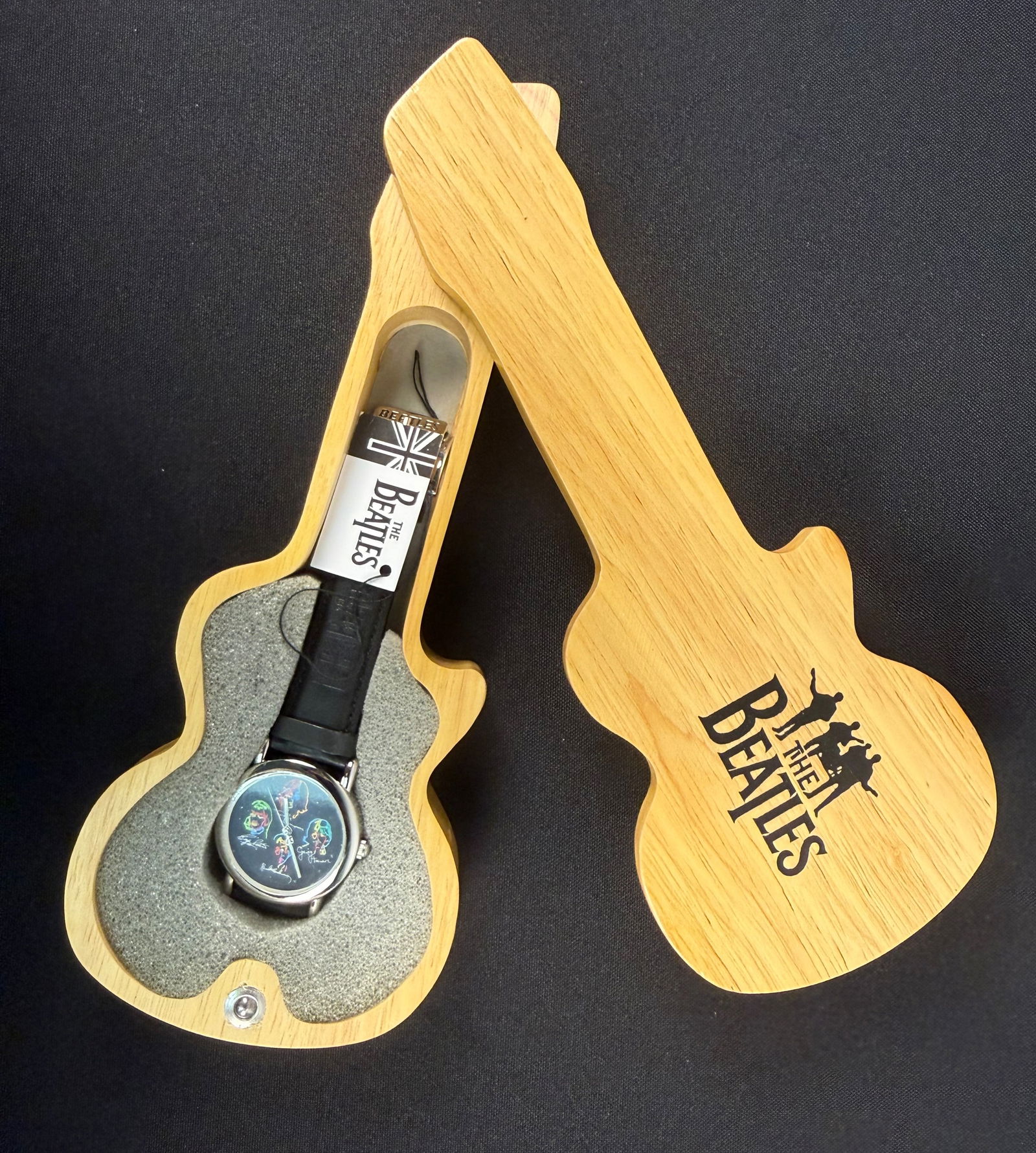 The Beatles Apple Corps Commemorative Wristwatch in Wooden Guitar Presentation Case: A Beatles Apple Corps commemorative wristwatch presented in a guitar-shaped wooden case. The watch features a silver-tone metal case and a black dial decorated with colorful sketches and facsimile sig