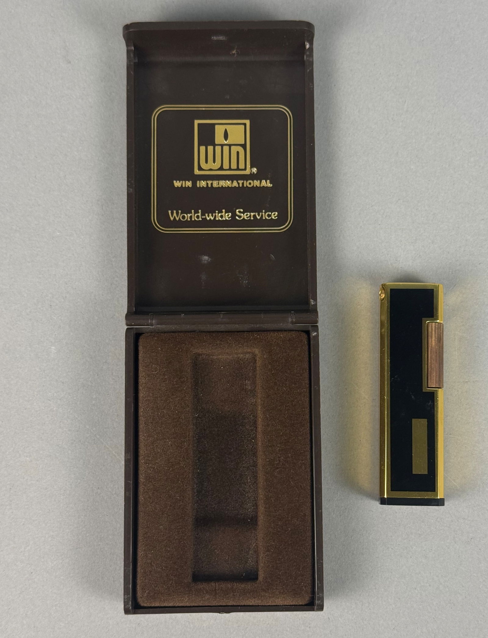 WIN International Butane Pocket Lighter, Japan: A WIN International butane pocket lighter manufactured in Japan. The lighter features a rectangular gold-toned metal body accented with black enamel side panels. The top includes a flip-cap mechanism,
