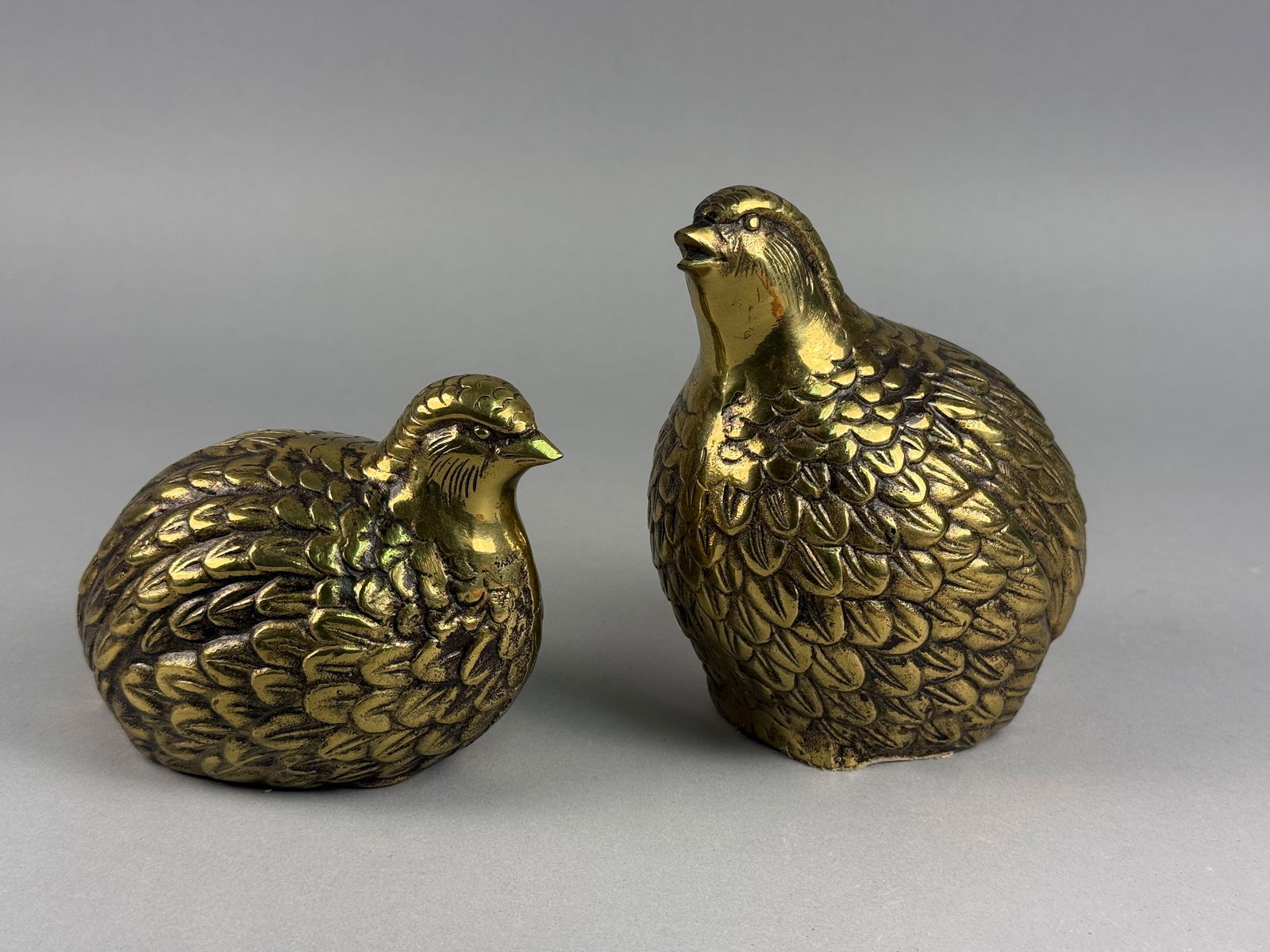 A Pair of Mid Century Modern Brass Quail Figurines: A pair of Mid Century Modern brass figurines depicting quail or partridges. The pieces are cast in a naturalistic style featuring detailed, textured feathering throughout the bodies. One figurine is m
