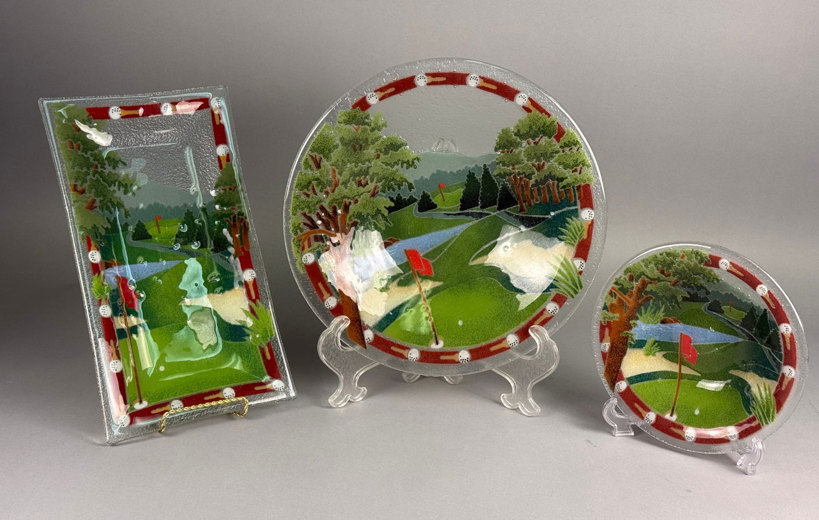 Peggy Karr Fused Art Glass Golf Course Landscape Collection Three Pieces: A collection of three fused art glass serving pieces by American artist Peggy Karr, featuring a matching stylized golf course landscape motif. The set comprises a rectangular tray, a large circular pl