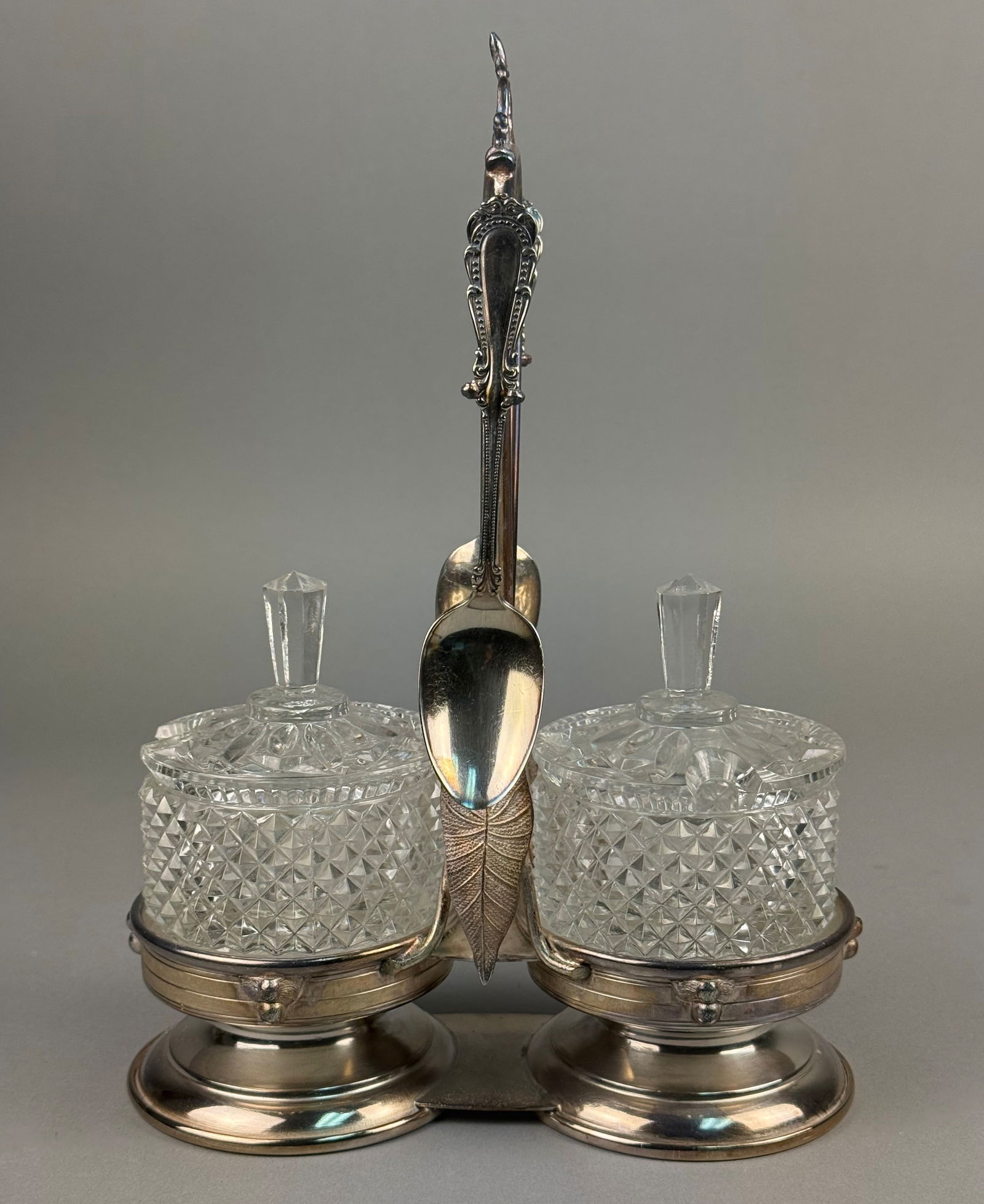 Rockford Silver Plate Co. Quadruple Plate Double Condiment Stand with Cut Glass Jars: A silver-plated double condiment or jam stand produced by the Rockford Silver Plate Co. The set includes a silver-plated frame, two cut-glass jars with matching faceted lids, and two silver-plated spo