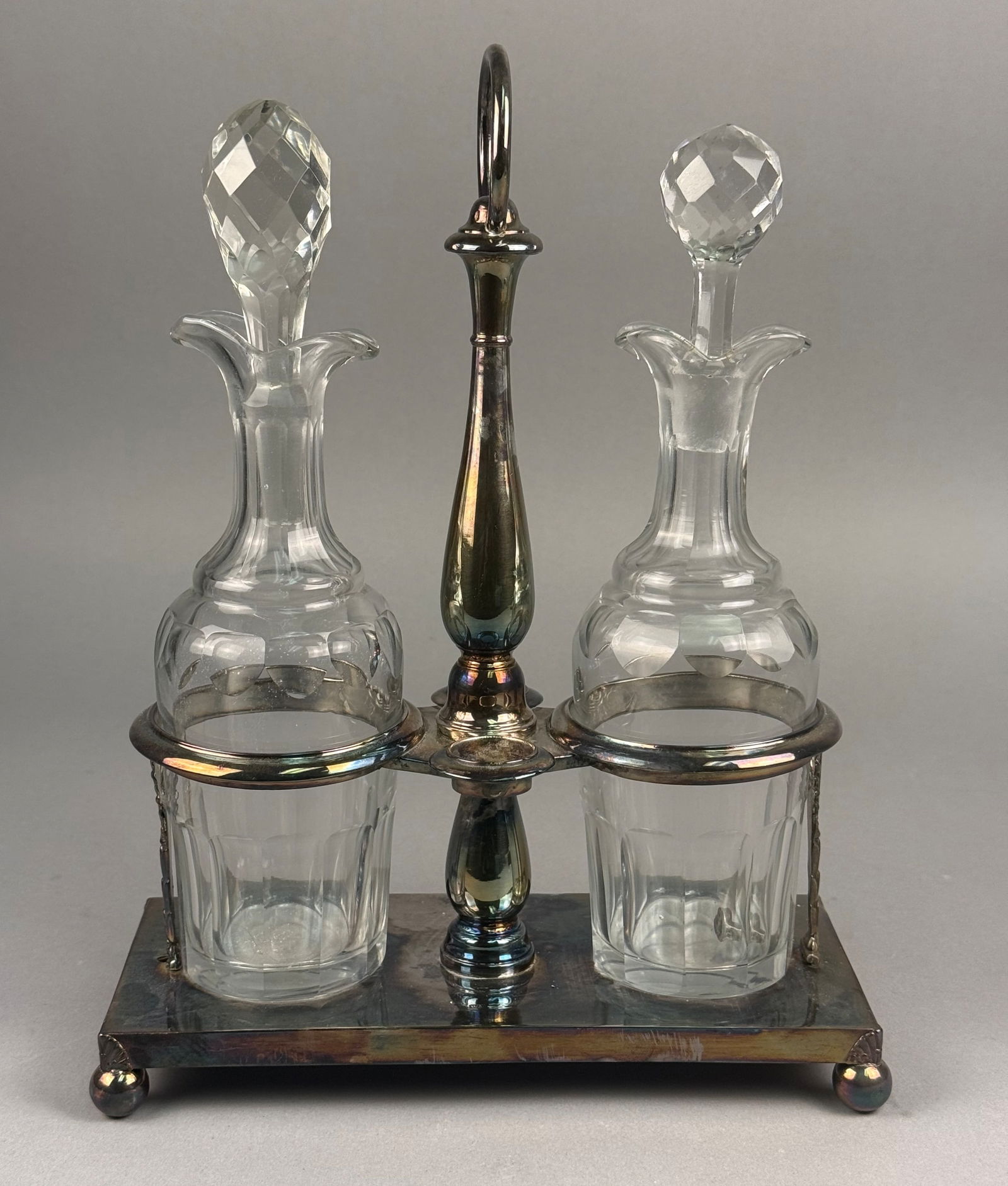 A Pair of Silver-Plated and Faceted Glass Cruets in a Stand: This silver-plated cruet set consists of two clear glass bottles with faceted stoppers housed in a rectangular metal stand. The stand features a central column topped with a ring-finial handle, accent