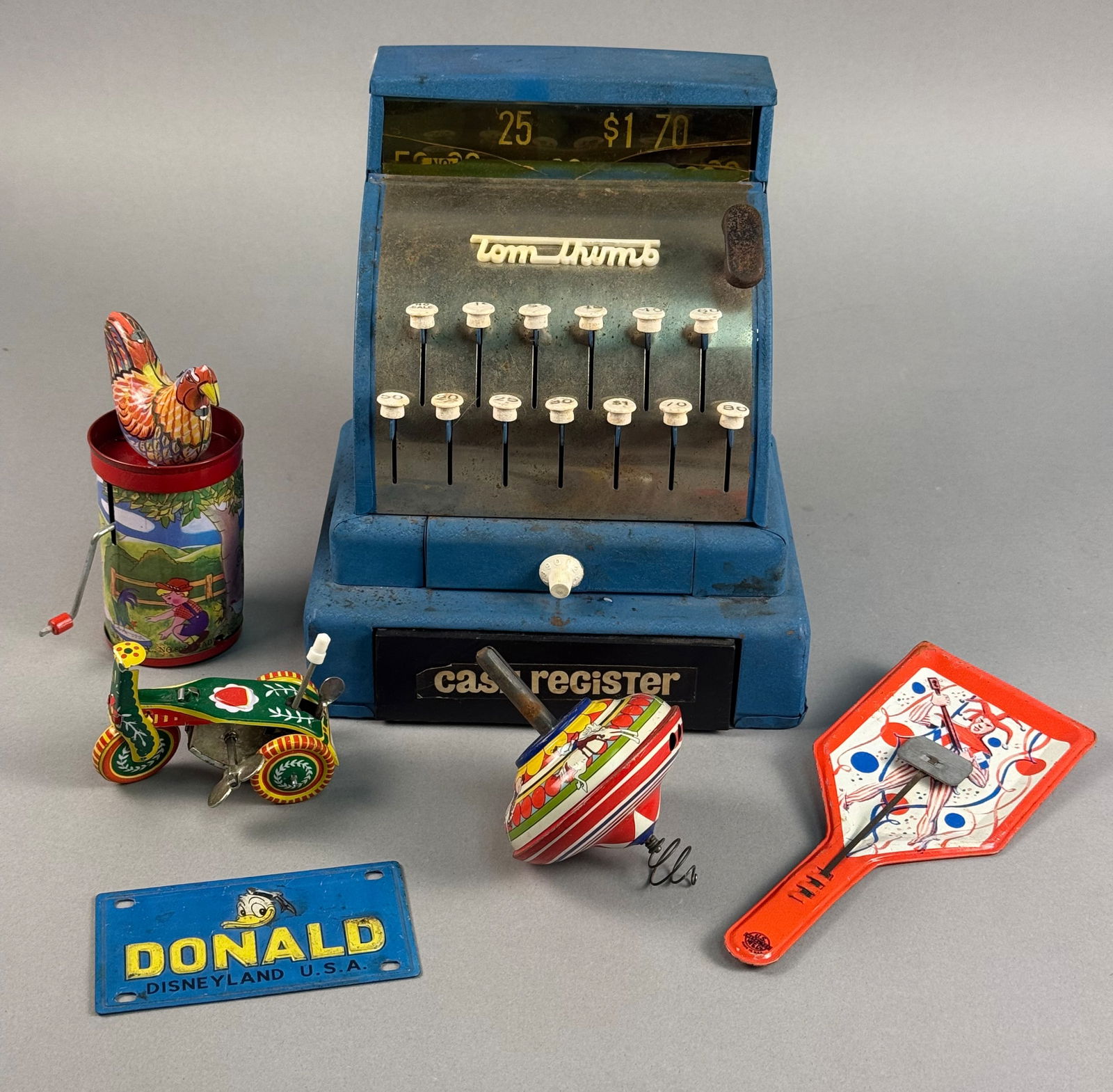 Vintage Tom Thumb Metal Cash Register and Assorted Tin Lithograph Toys, c. 1950s-1970s: A diverse group of mid-century and later vintage toys. The primary piece is a blue Tom Thumb brand toy cash register, constructed of pressed steel with a crinkle-finish paint. It features a series of