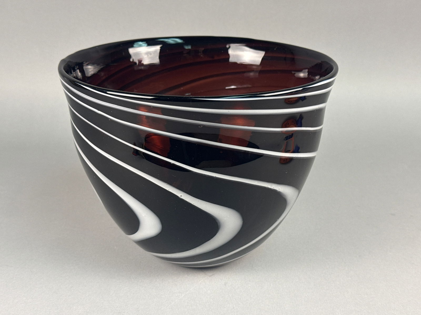 Large Hand-Blown Amethyst and White Latticino Spiral Art Glass Bowl: This sophisticated hand-blown art glass vessel features a deep amethyst glass body, so saturated it appears nearly black in ambient light, revealing a translucent reddish-purple glow when backlit. The