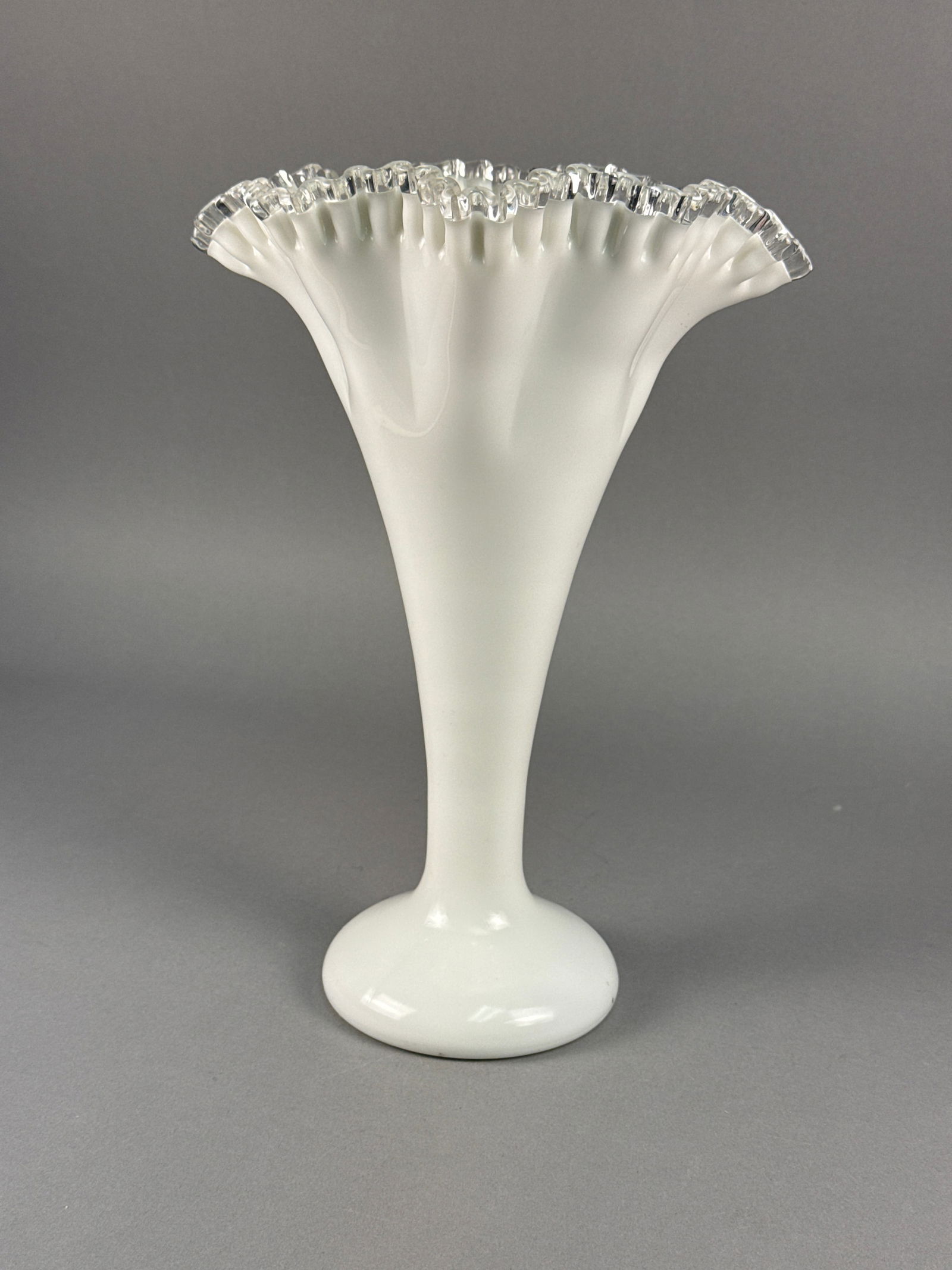 Milk Glass Fluted Trumpet Vase: The piece is composed of opaque white glass featuring a flared, ruffled rim with a clear glass crimped edge. The body tapers down to a smooth, circular pedestal base. This style of glass was character