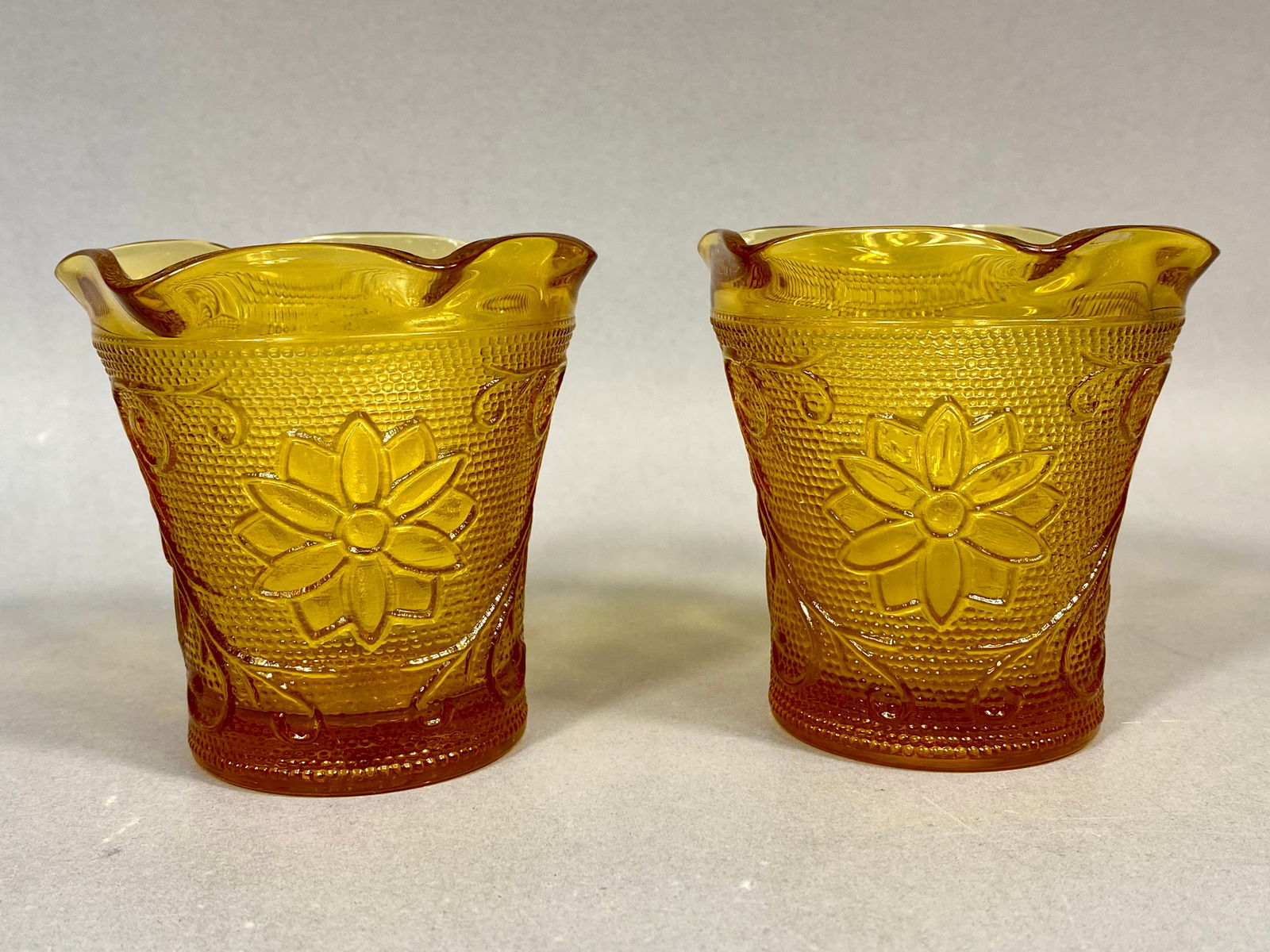 Tiara Exclusives, Amber Pressed Glass, A Pair of Votive Cups: This pair of translucent amber glass votive cups was produced for Tiara Exclusives in the Sandwich pattern. The design features mold-pressed embossed floral daisy and scroll motifs set against a stipp