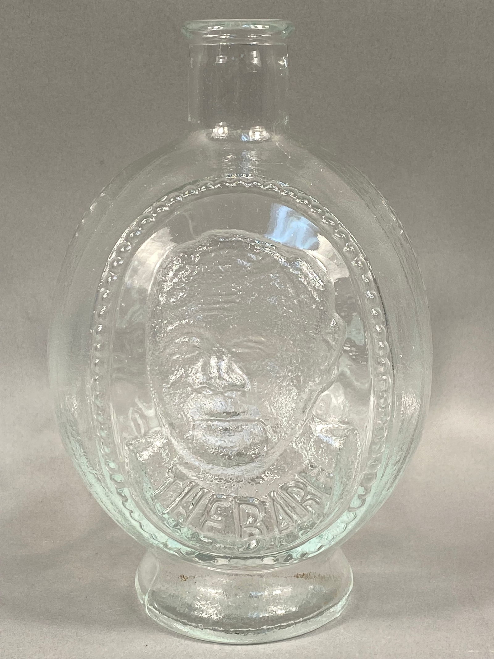 Tiara Exclusives Molded Clear Glass Commemorative Sports Heroes Bottle: A vintage clear pressed glass commemorative bottle produced for Tiara Exclusives, featuring high-relief molded portraits of legendary American athletes. The flattened oval body displays three distinct